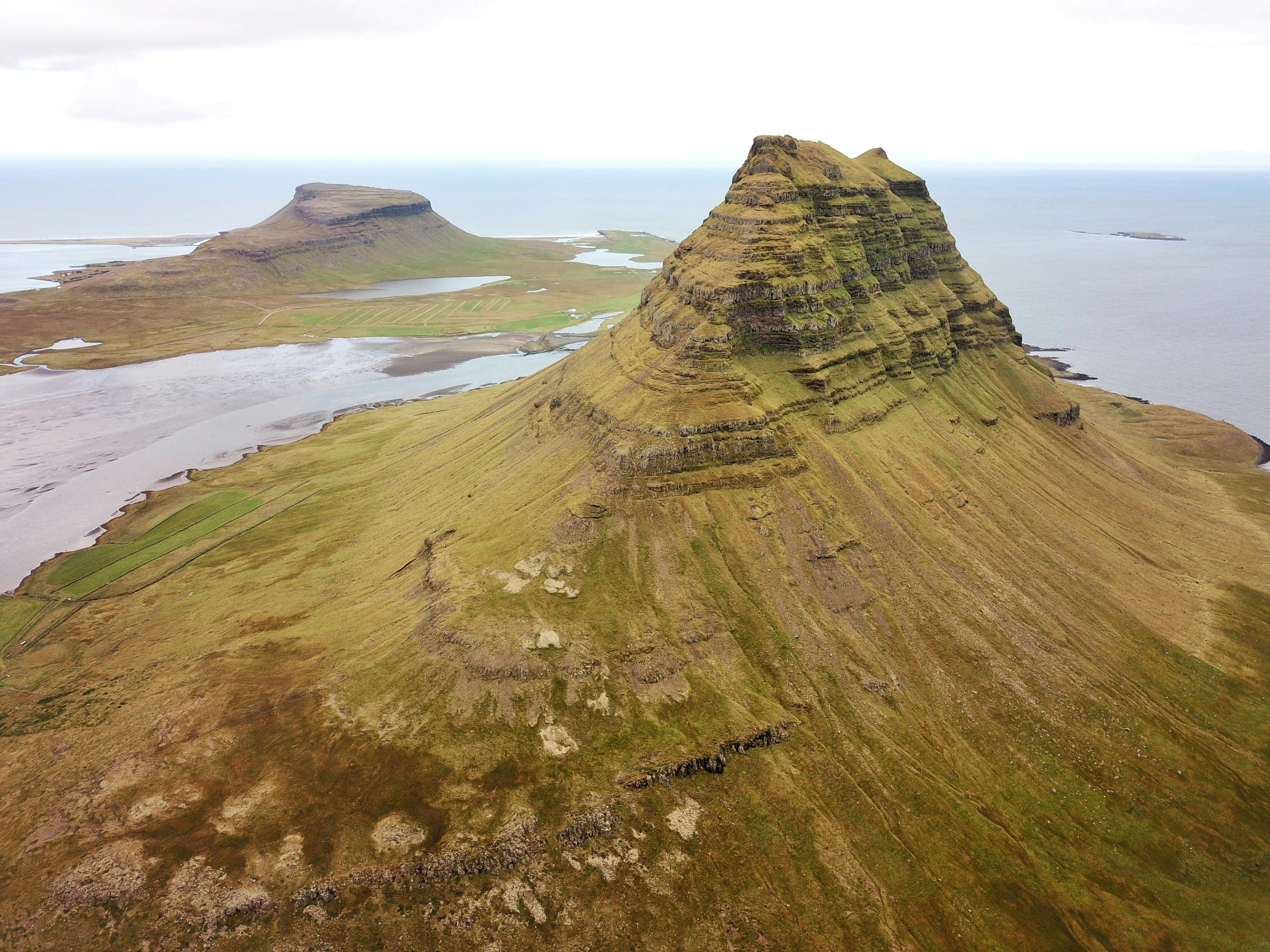A prominent green conical mountain with layered rock formations stands by the sea, with a flatter mountain and grassy land under a cloudy sky.