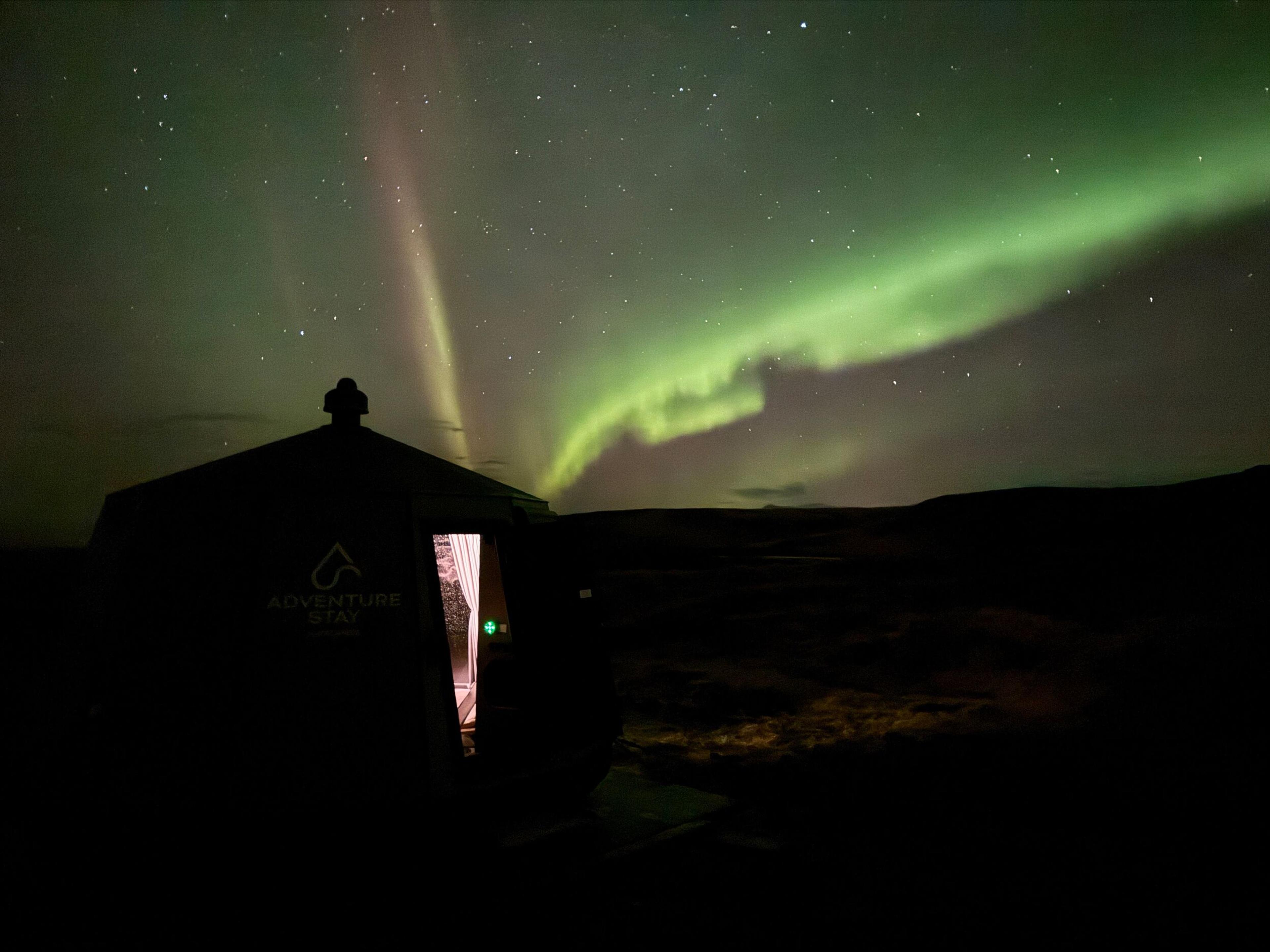 A dark 'Adventure Stay' cabin with an open door beneath a starry sky illuminated by vibrant green and purple Northern Lights.