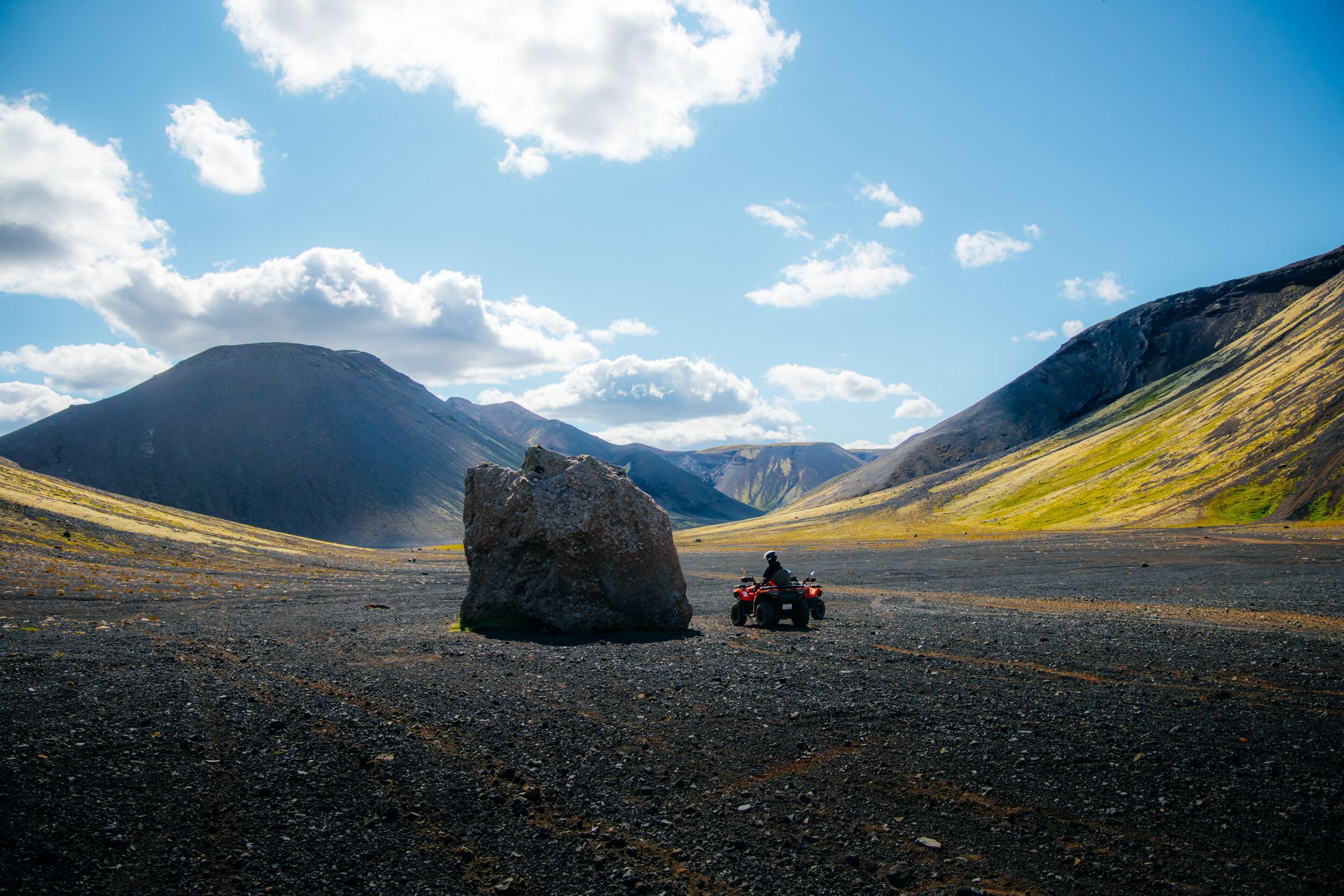 A person rides an ATV on a dark, rocky plain with a large boulder and mountains under a blue sky.