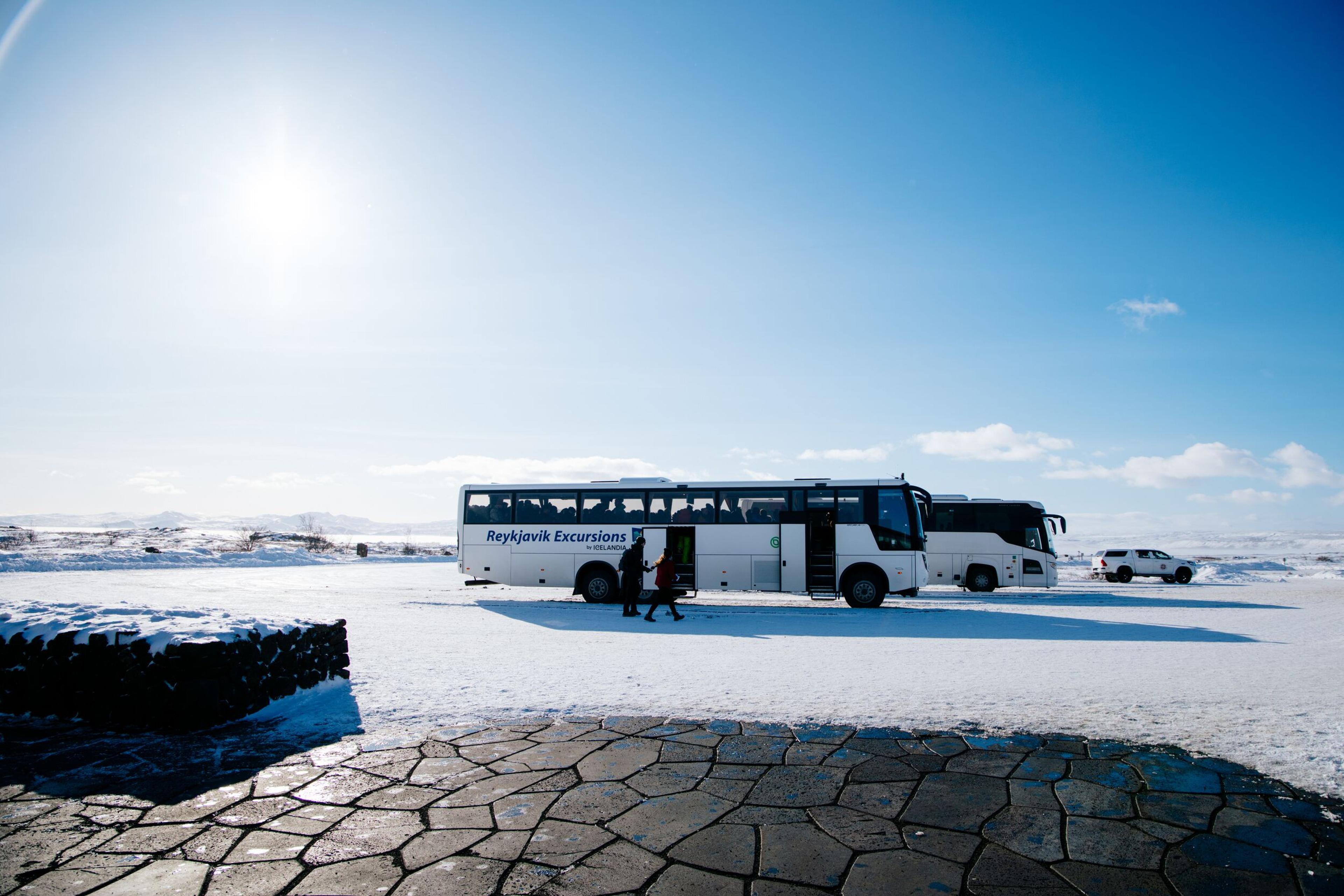 White 'Reykjavík Excursions' tour buses parked in a snow-covered lot under a bright blue sky, with passengers boarding the front bus.