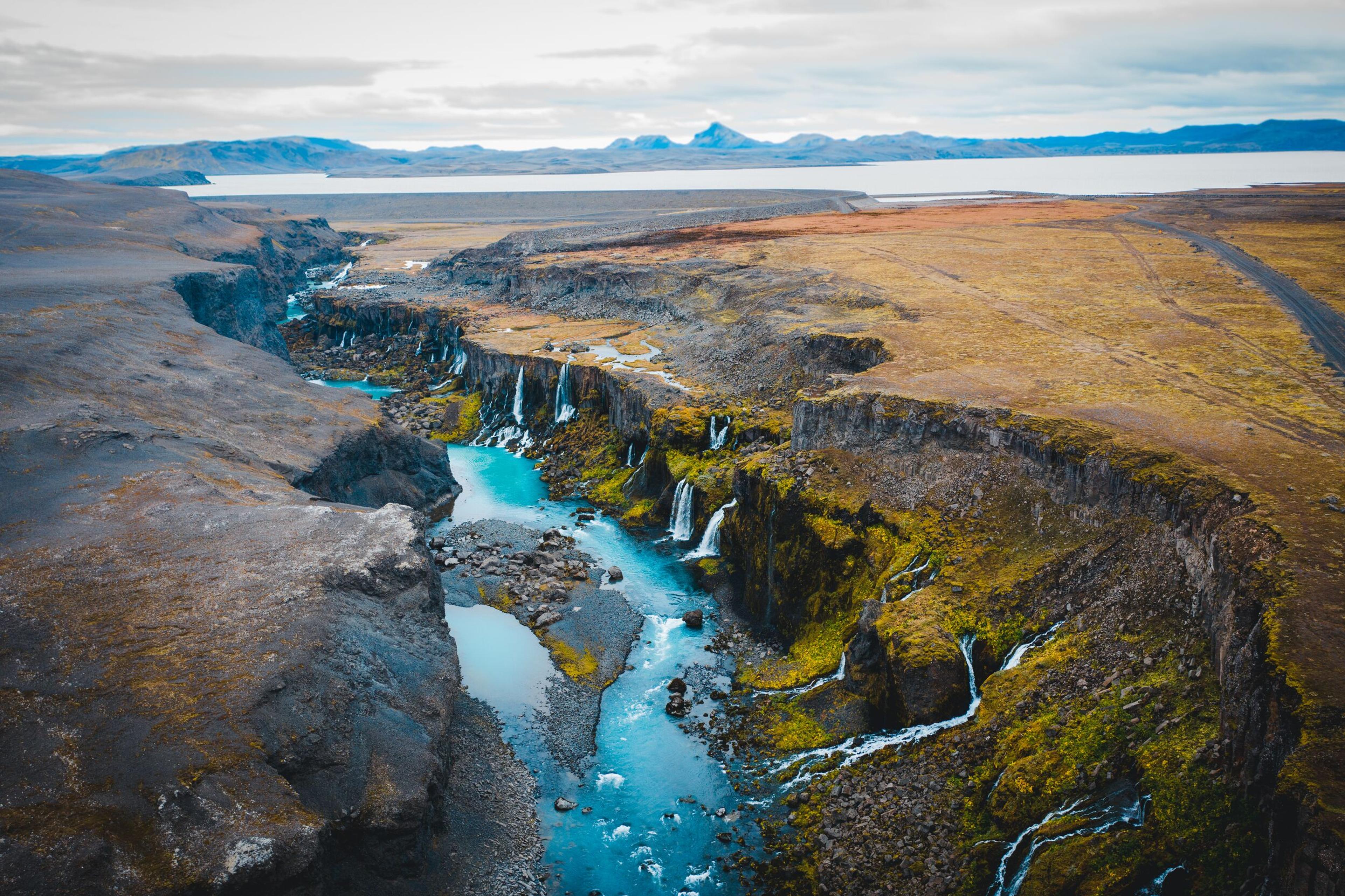 A turquoise river winds through a deep canyon with numerous waterfalls cascading down its mossy walls, surrounded by barren land and distant mountains and a lake.