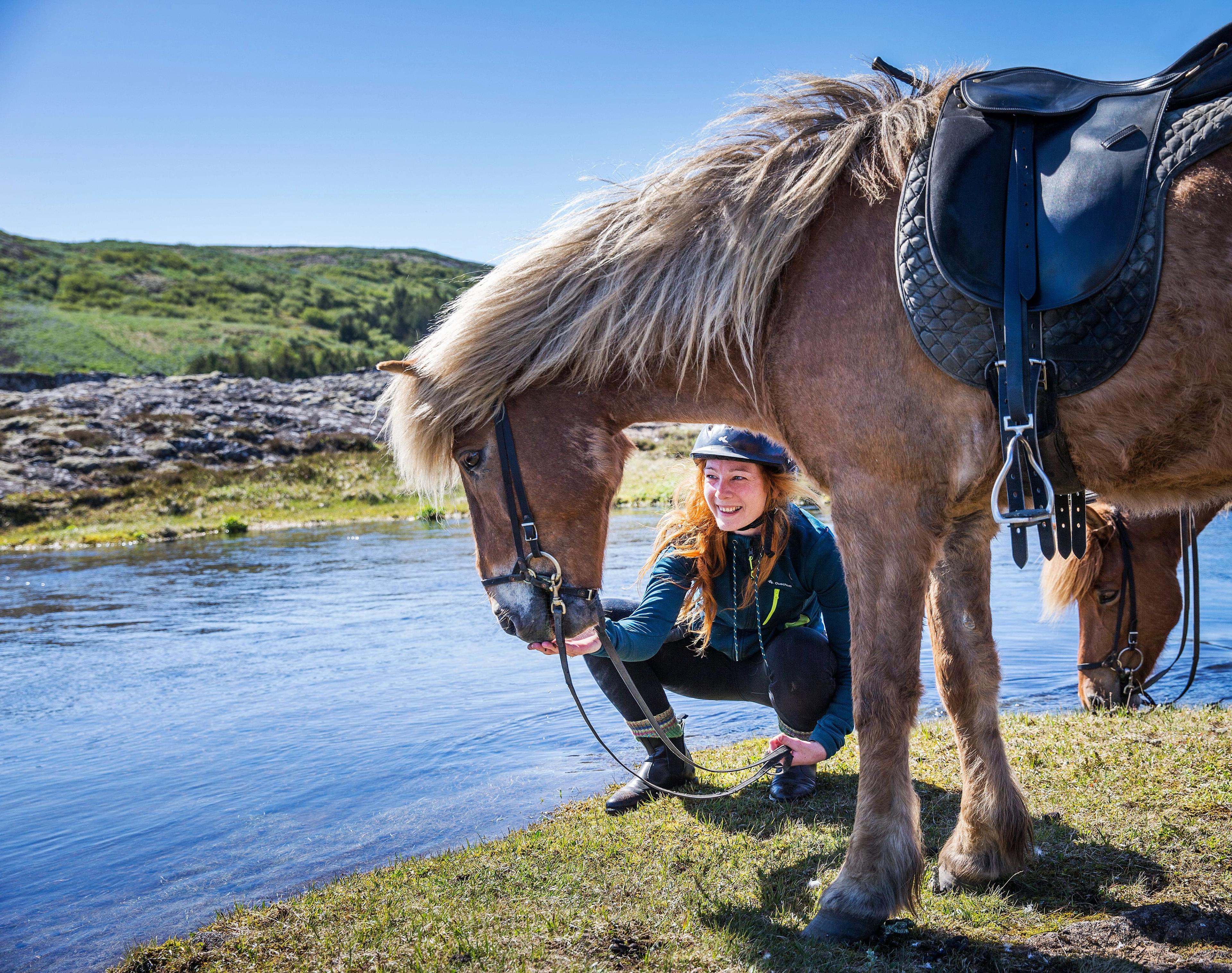 A smiling woman kneels beside a saddled horse by a river, with another horse drinking in the background.