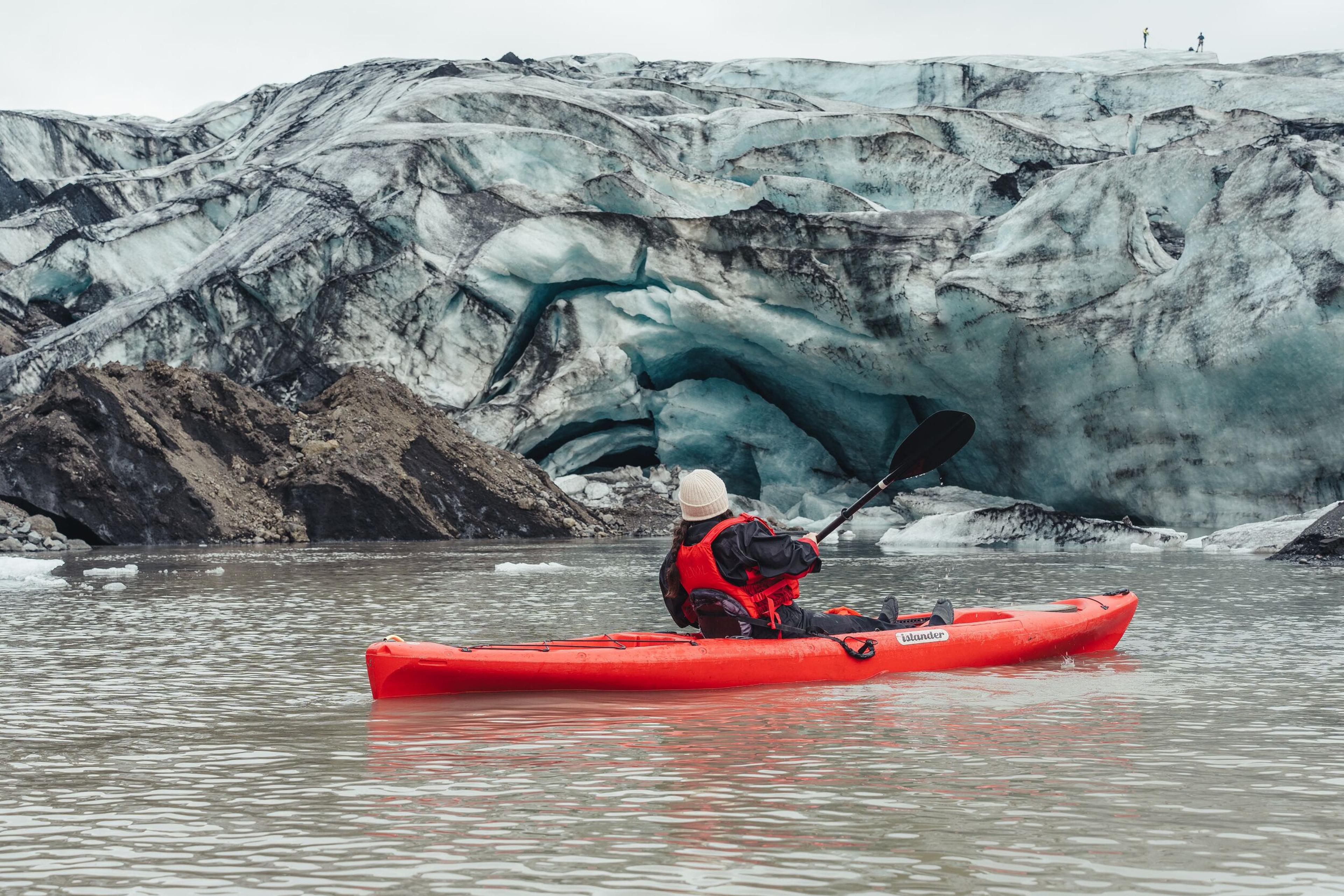 A person in a red kayak paddles on a glacial lake with a large glacier in the background.