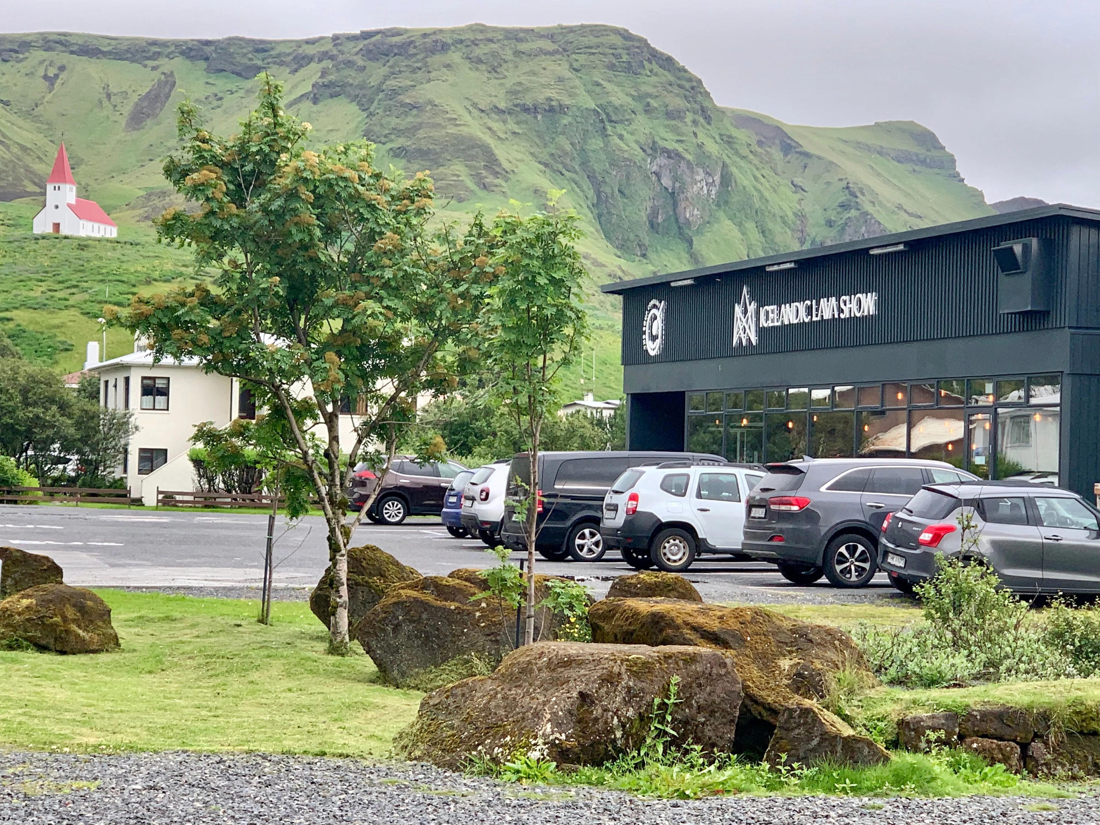 A black building for the Icelandic Lava Show, a parking lot with cars, and a white church with a red roof on a green hill, surrounded by mountains and mossy rocks.