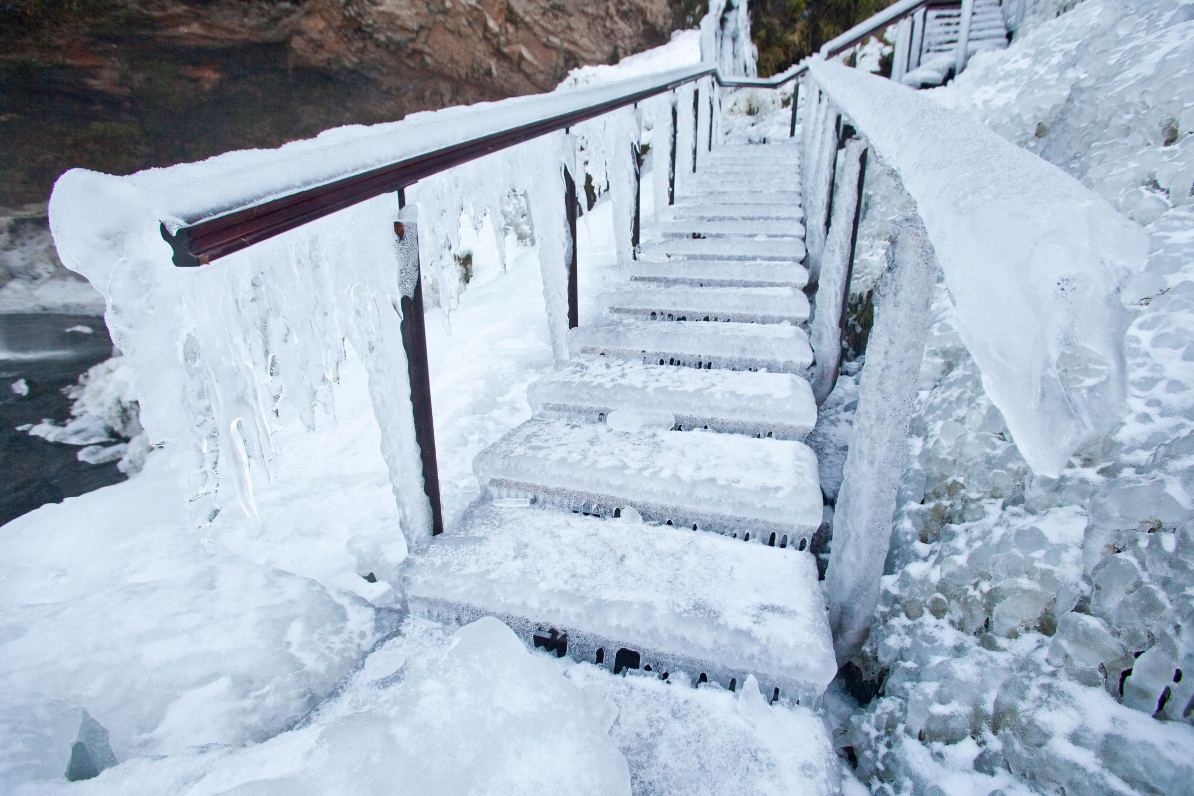 A stairway leading toward Seljalandsfoss waterfall is entirely coated in thick ice, with icicles hanging from the railings and steps.