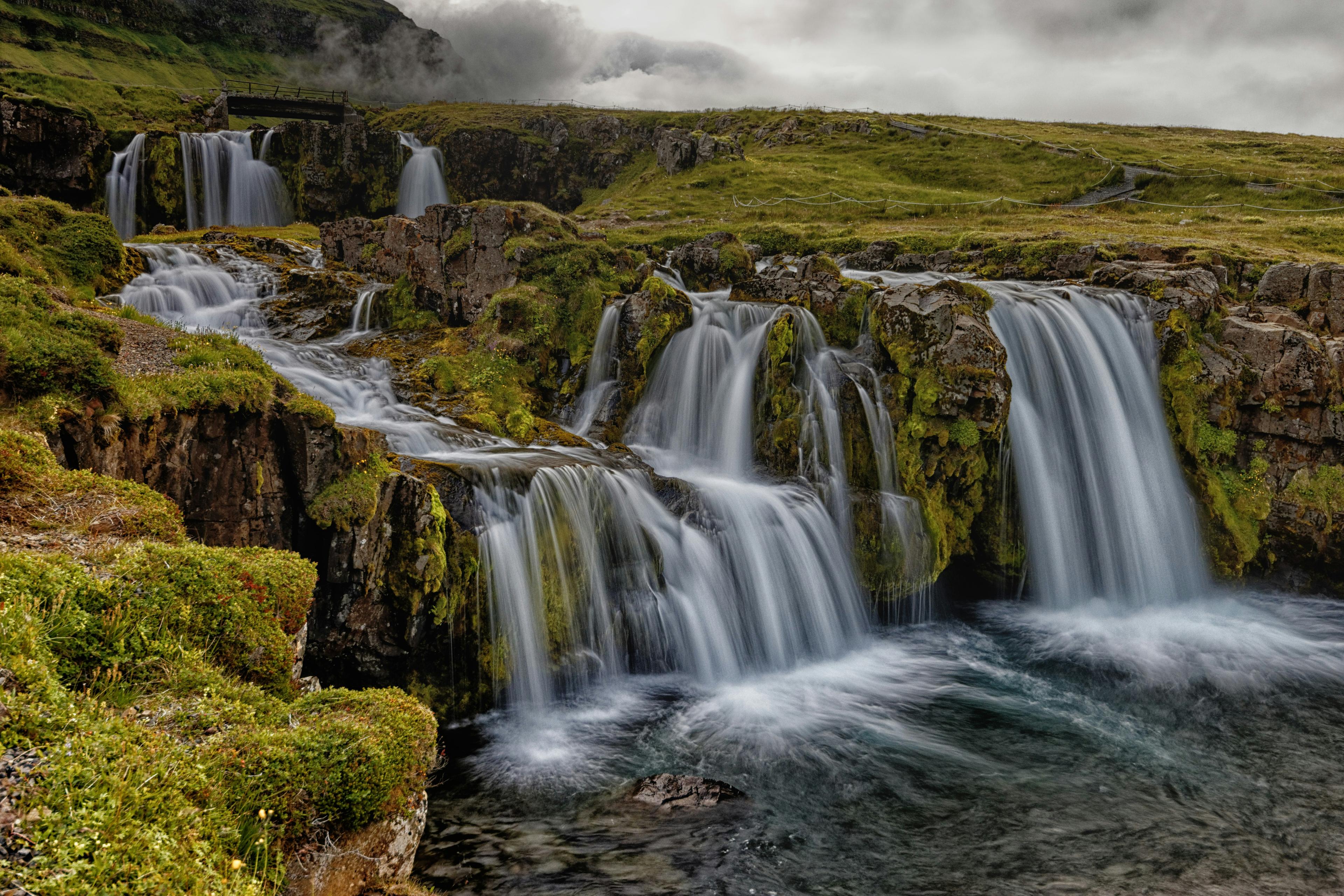 A breathtaking view of Kirkjufellsfoss waterfall in Iceland, cascading over moss-covered rocks into a crystal-clear pool, surrounded by lush green landscape and misty clouds.