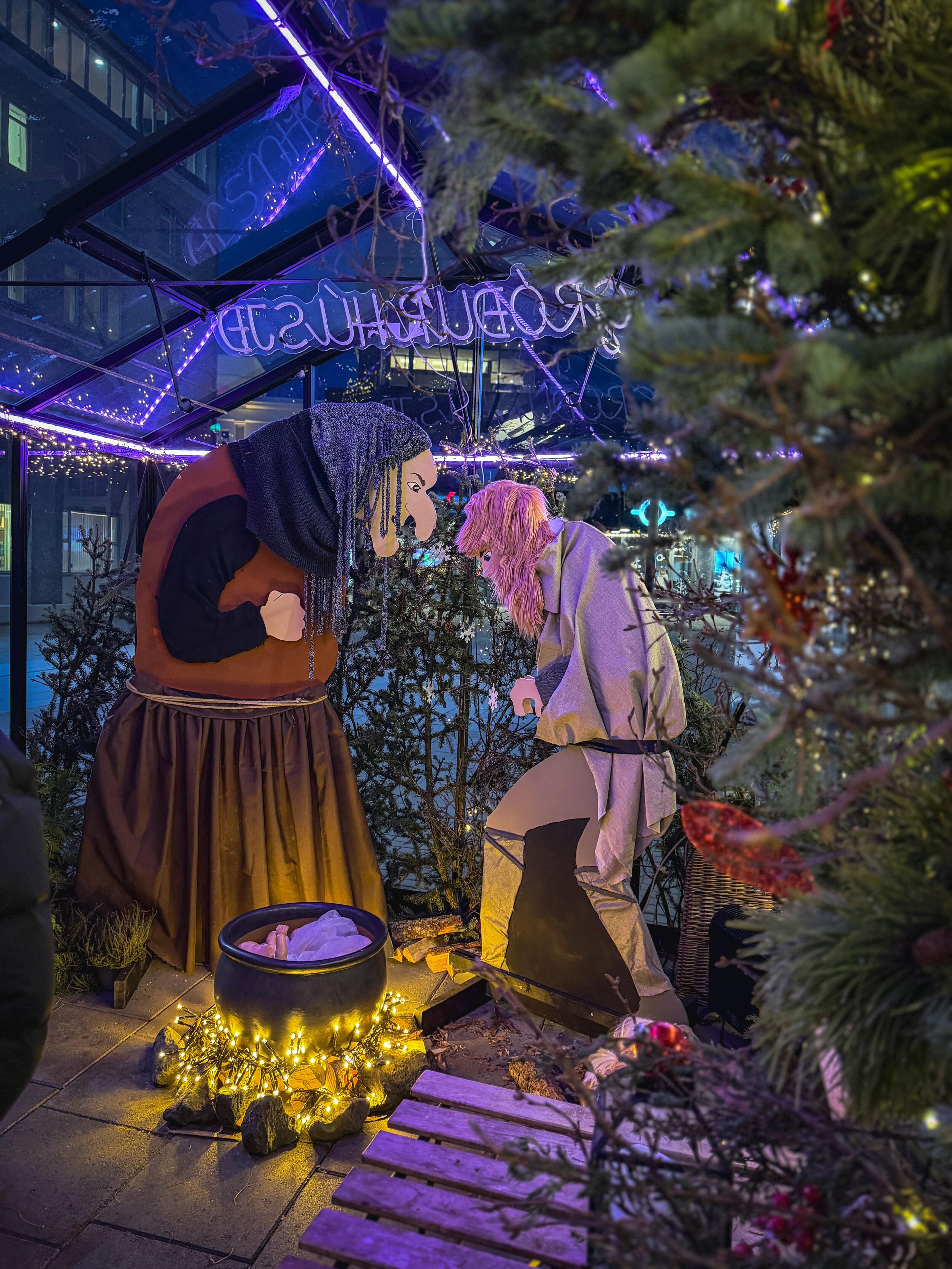 A fairytale display with an old woman and a younger person next to a glowing cauldron, under a glass roof with purple neon lights reflecting "Hans Christian Andersen's".