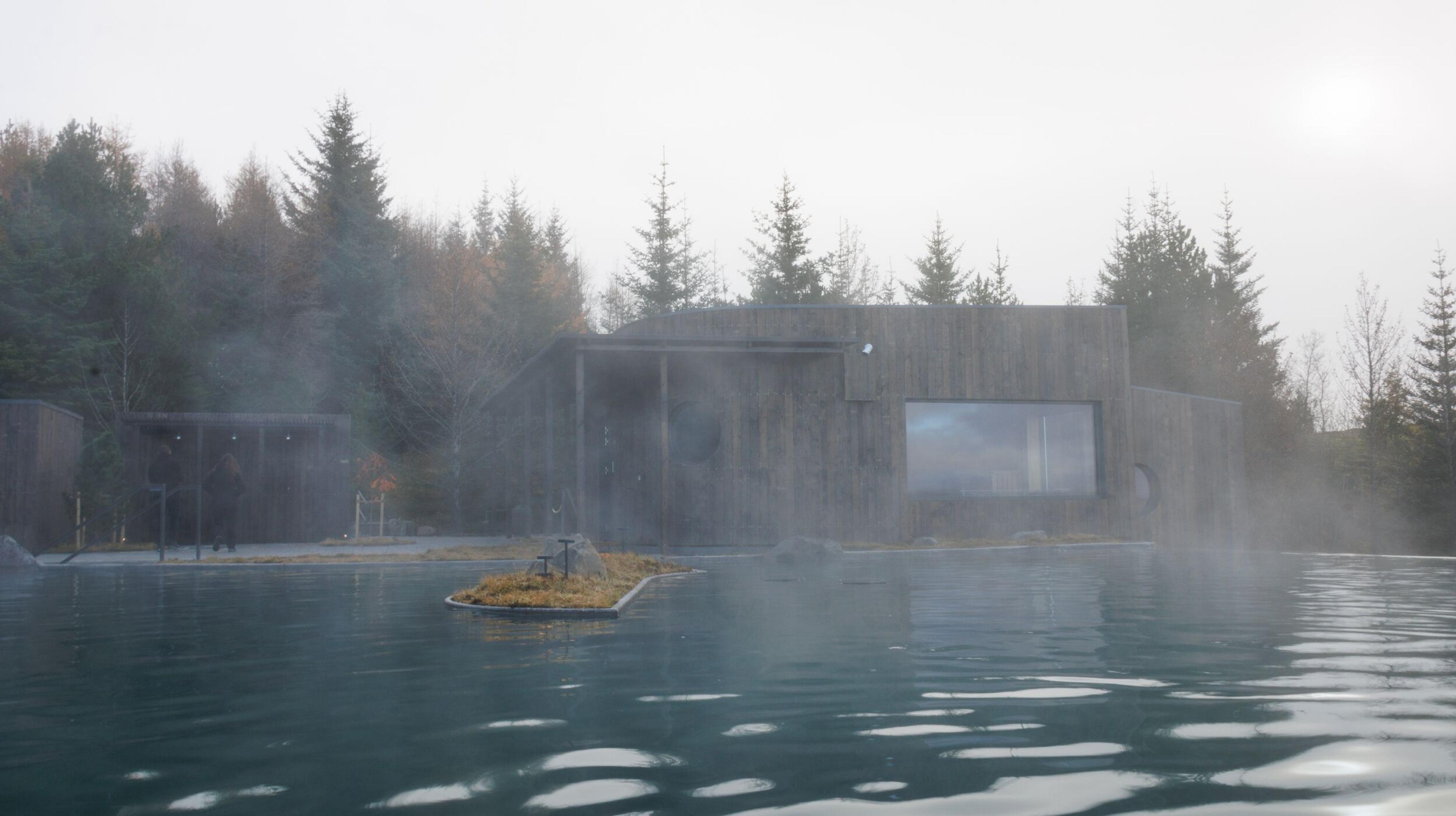 A steaming geothermal pool in front of a modern wooden building and pine forest.