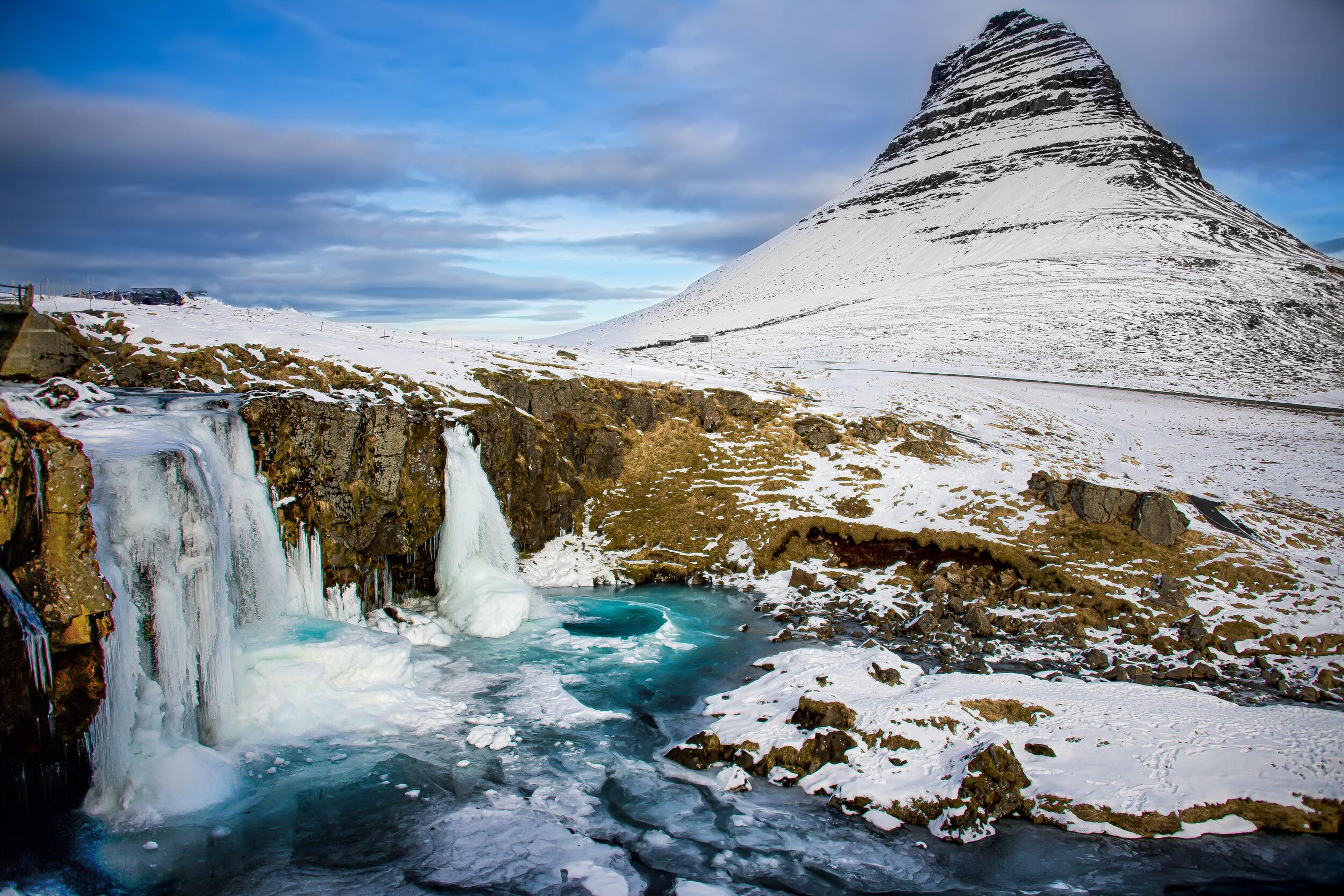 Snow-covered Kirkjufell mountain overlooks a partially frozen Kirkjufellsfoss waterfall and a turquoise river.