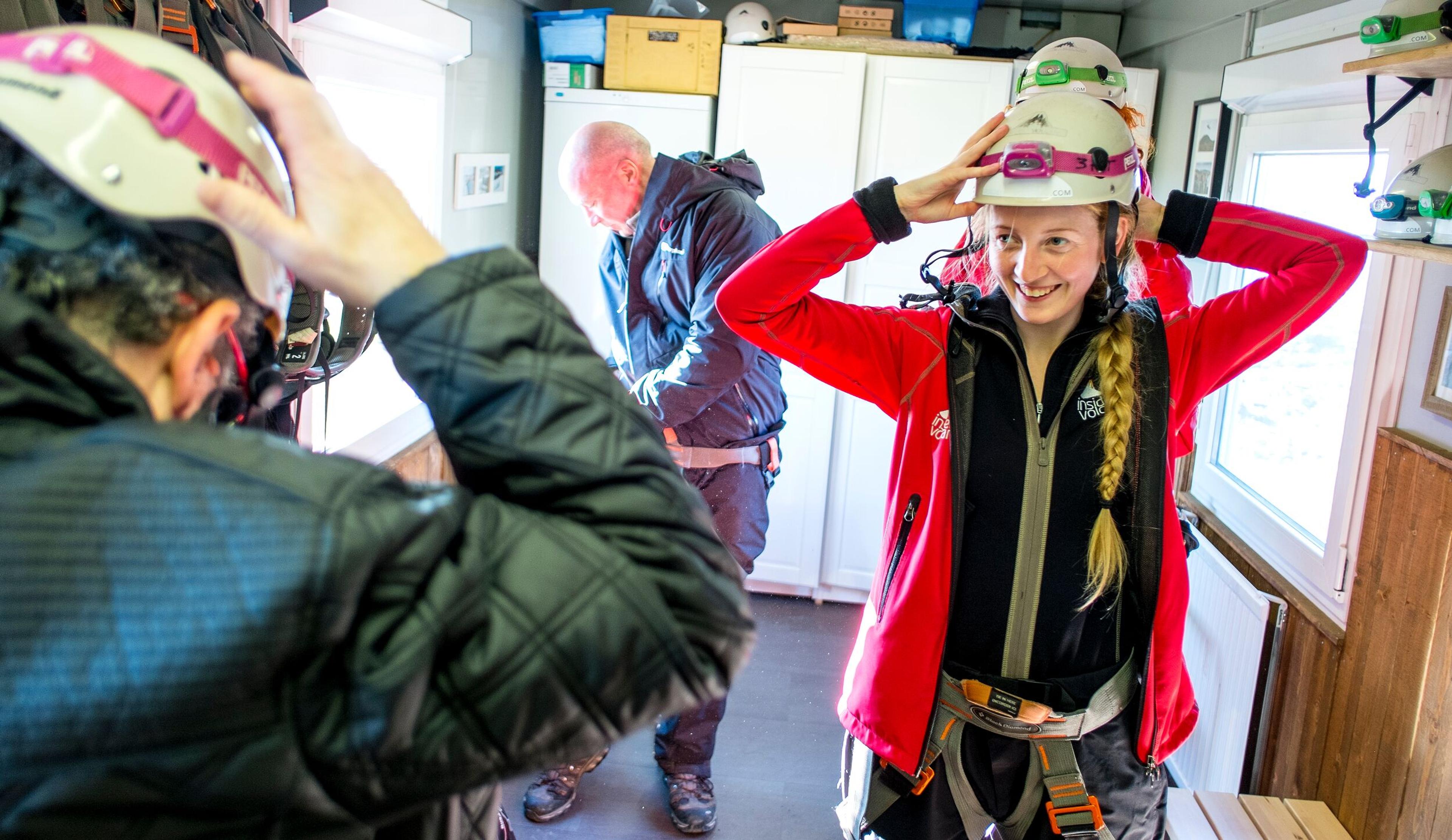 People in an indoor room trying on helmets and harnesses. A woman in a red jacket smiles while wearing two helmets.