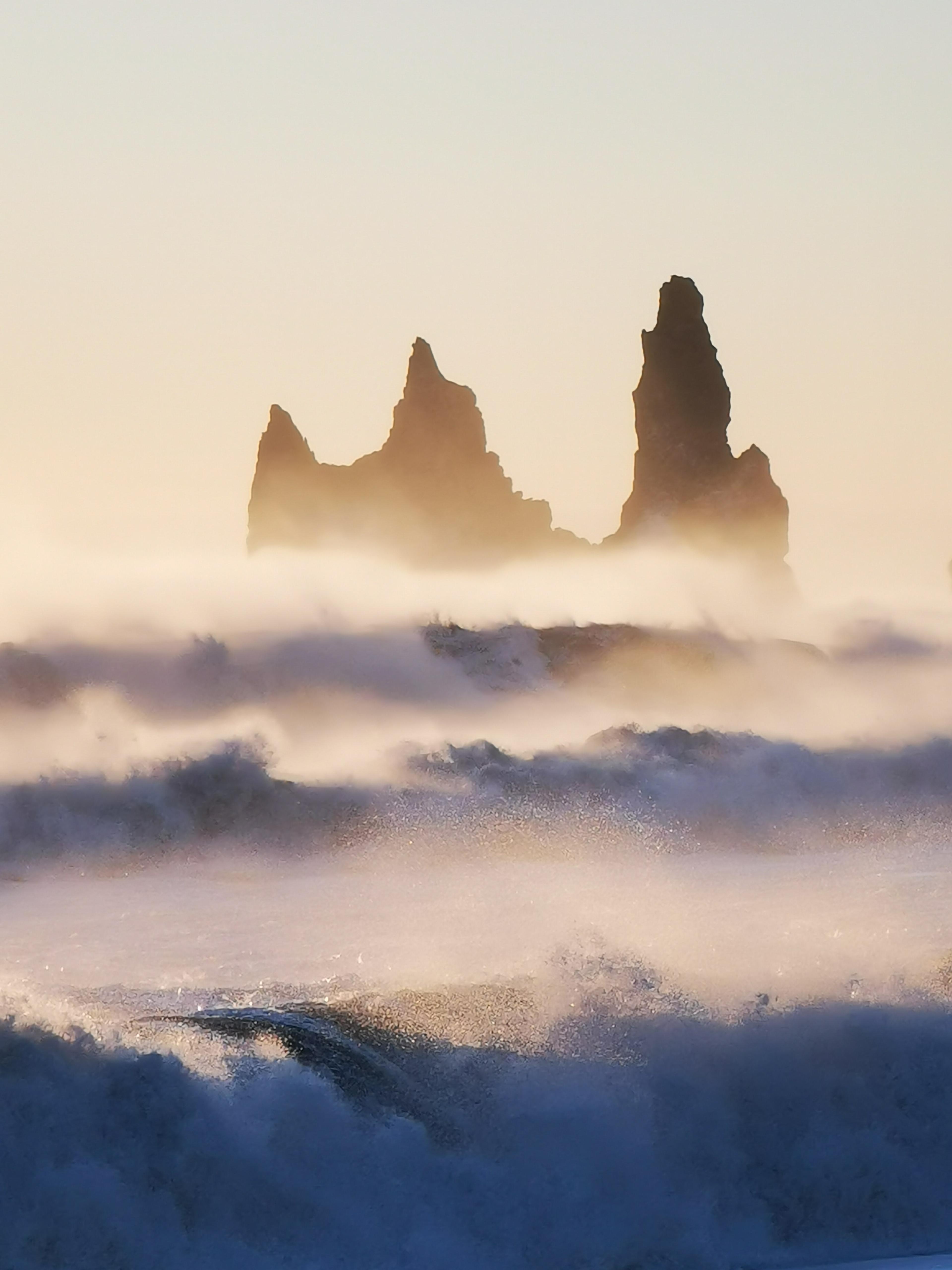 Massive waves crashing onto the black sands of Reynisfjara beach, with the iconic Reynisdrangar sea stacks looming in the misty background.