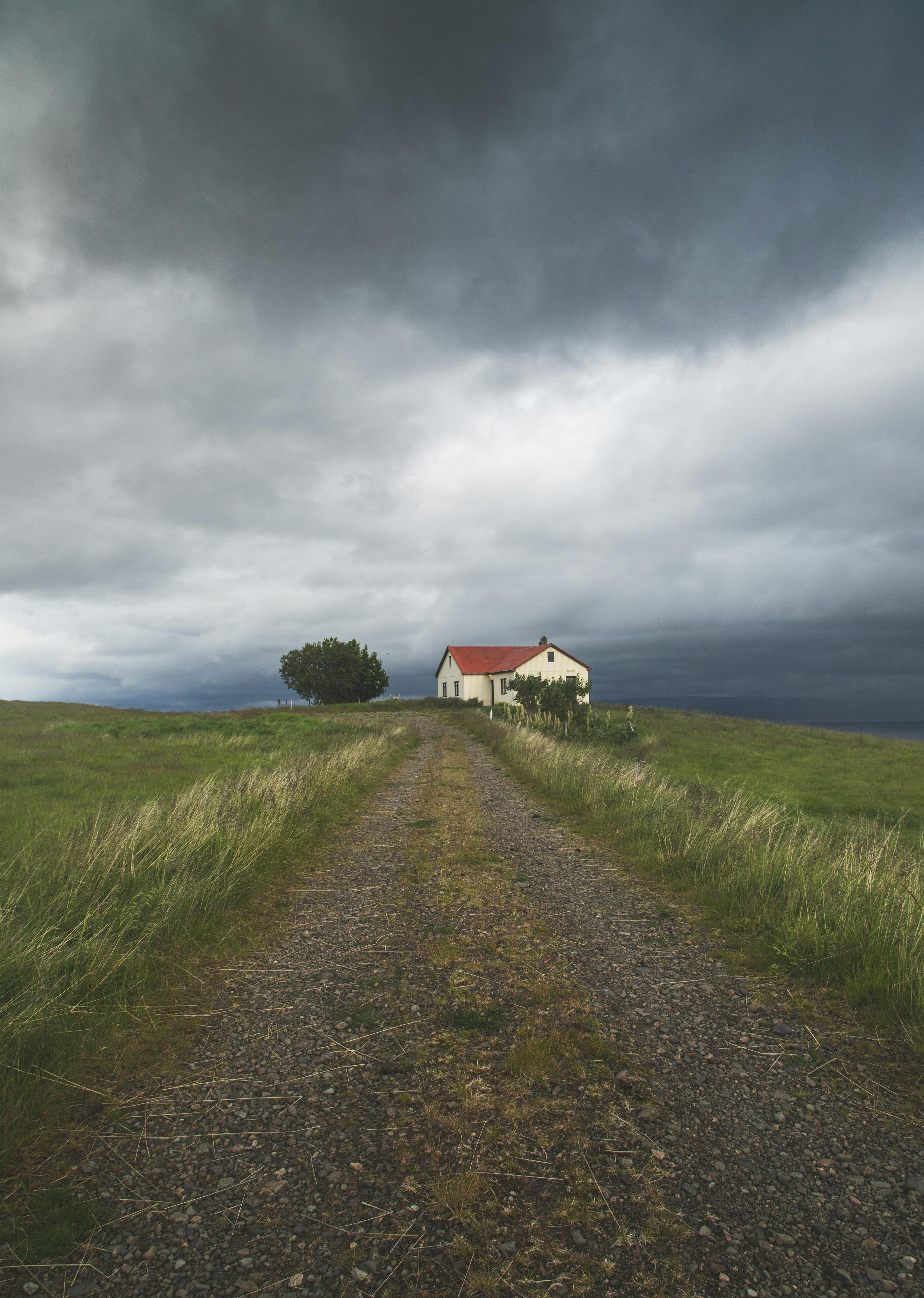 A lonely house with a red roof sits atop a grassy hill, framed by a winding dirt path and a dramatic stormy sky in the background.