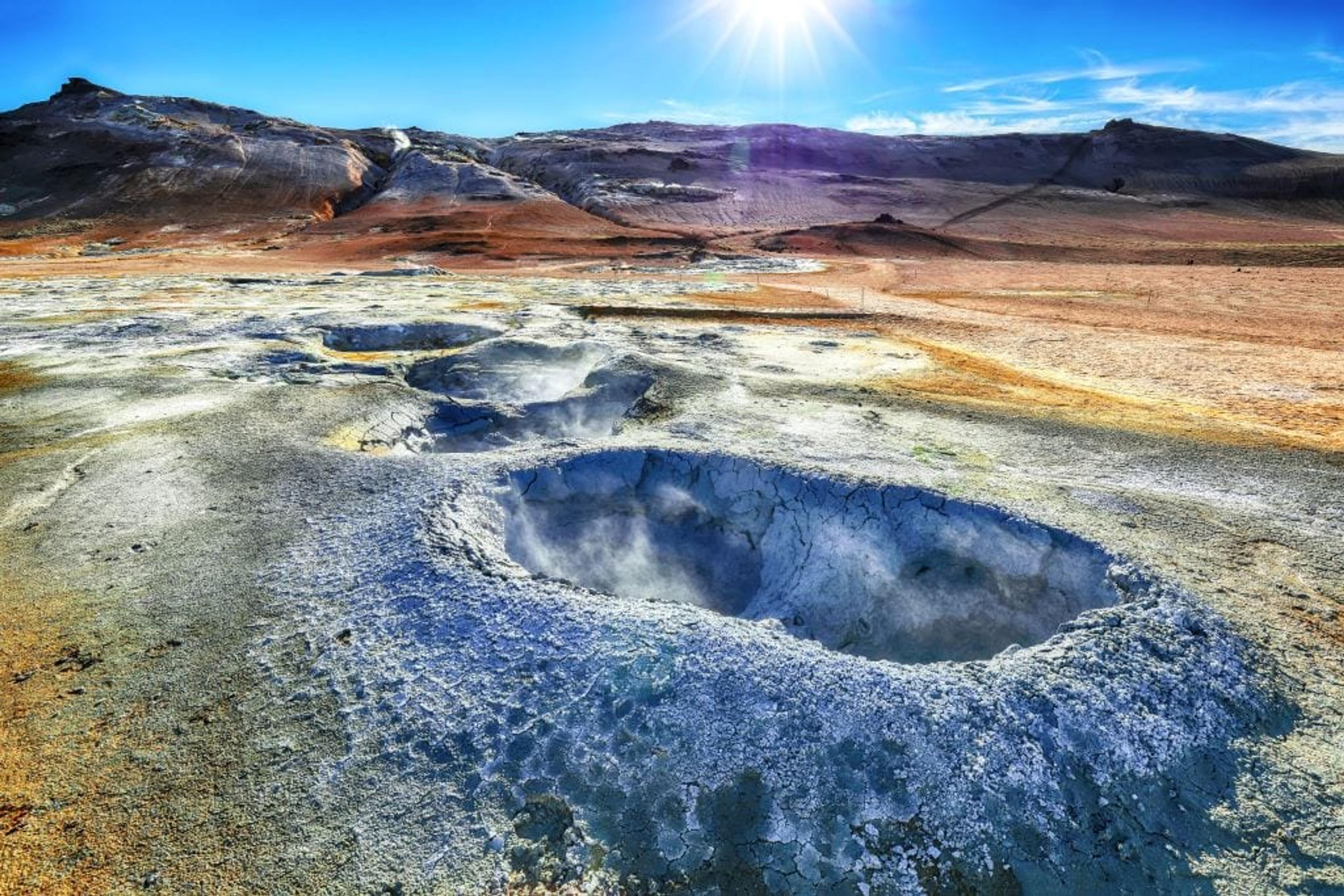 Geothermal field with steaming vents, barren hills, and a bright sun in a blue sky.