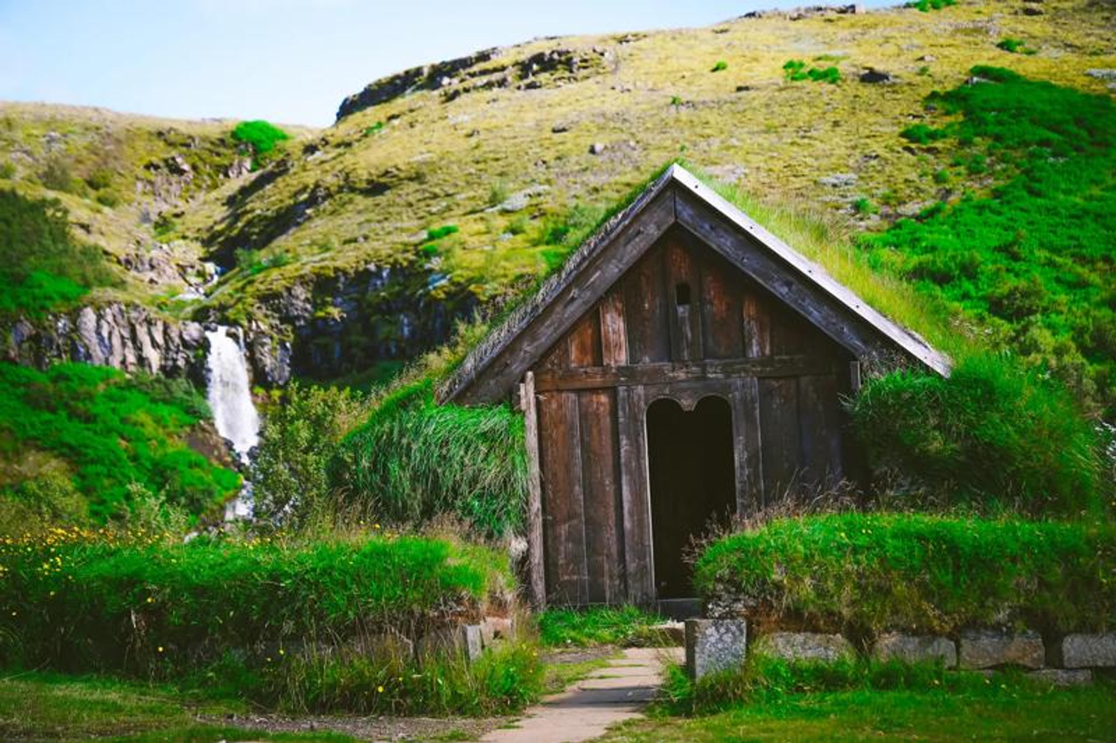 A rustic wooden house with a grass roof stands in a lush green valley with a waterfall.