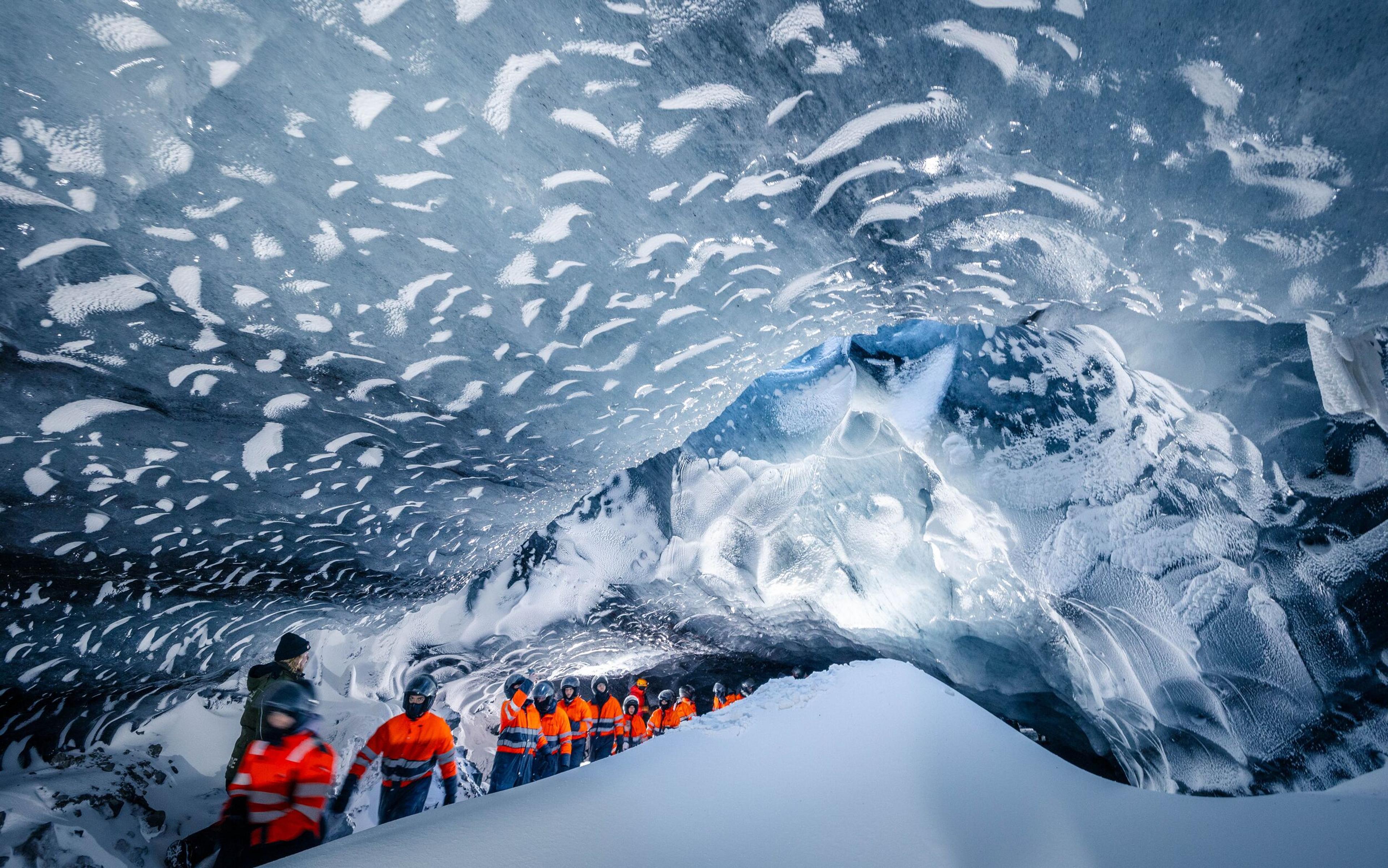 Group of tourists in orange jackets exploring a stunning blue ice cave with intricate ceiling patterns.