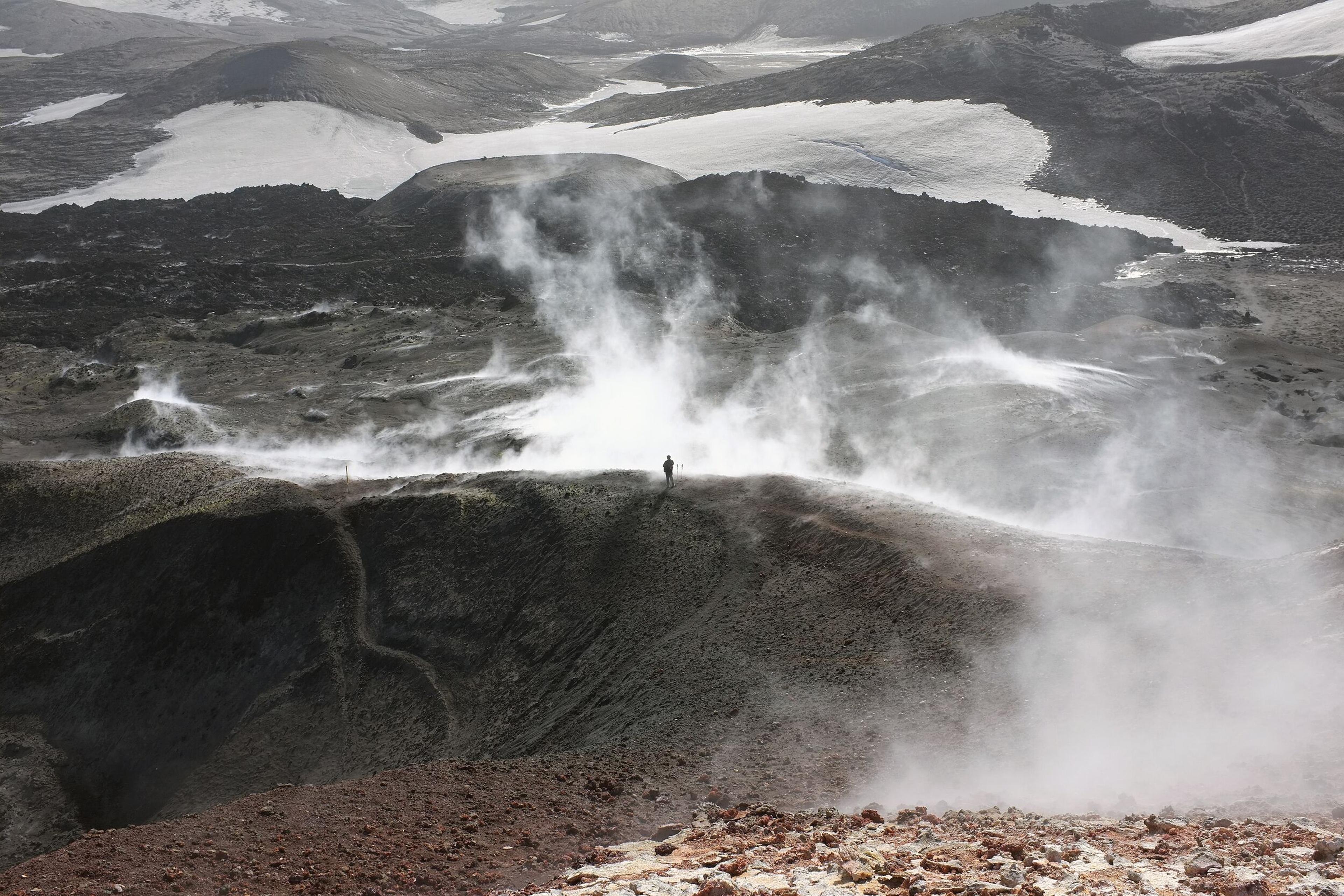 A lone hiker standing near steaming geothermal vents on Fimmvörðuháls, surrounded by rugged volcanic terrain and patches of snow.