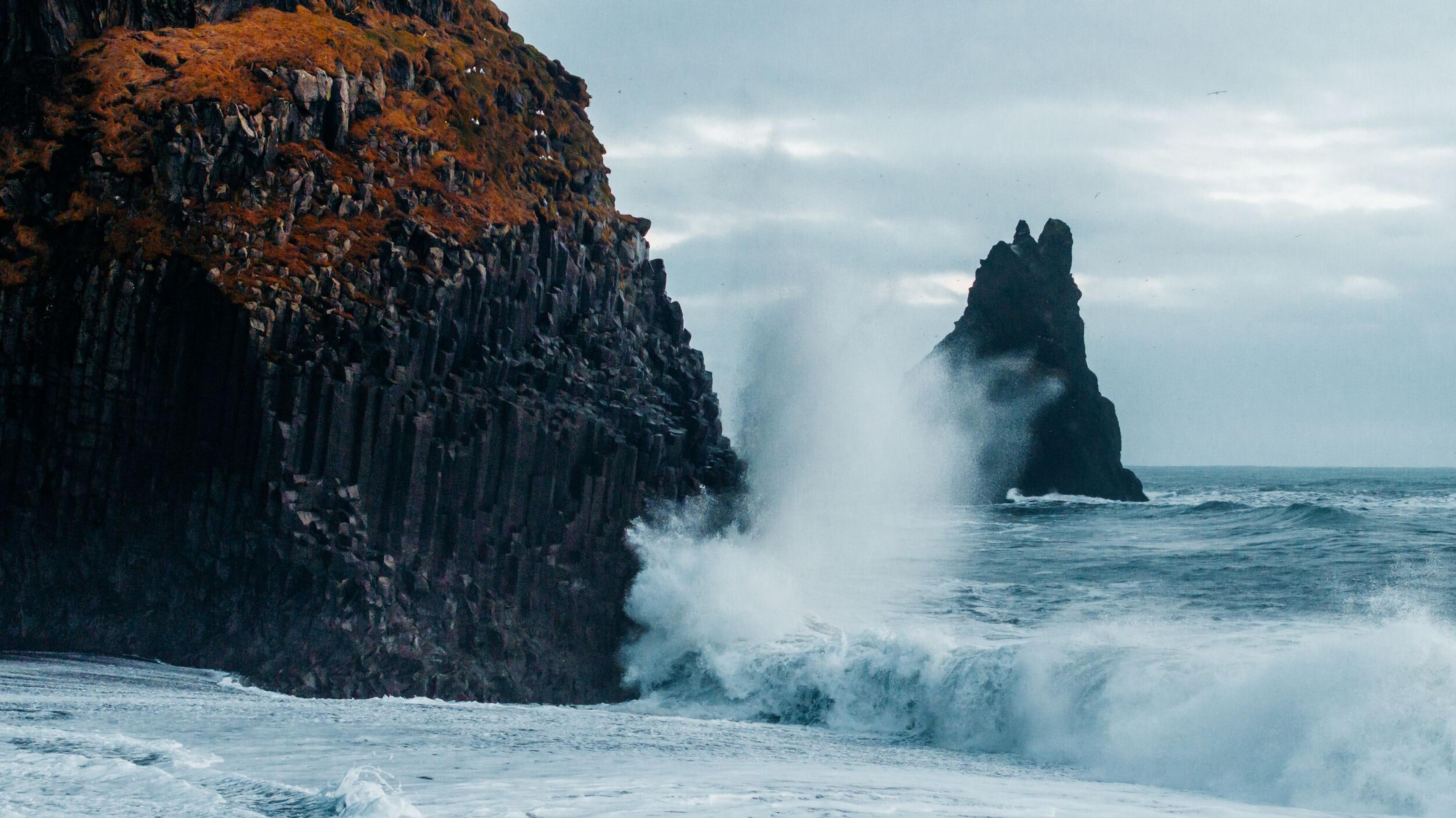 Powerful waves crash against a columnar basalt cliff, with a sea stack in the misty distance under an overcast sky.