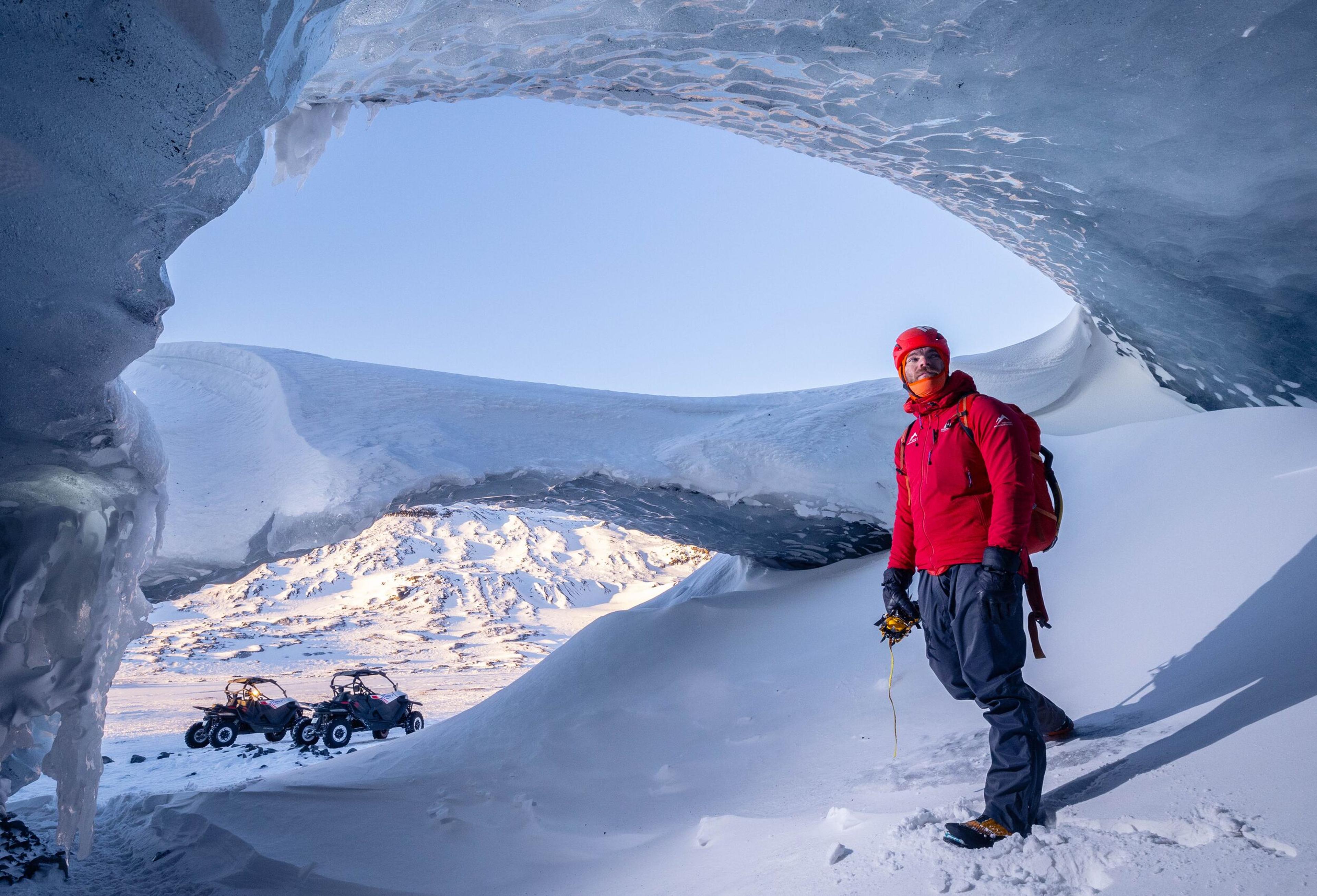 A person dressed in red winter gear stands inside the Askur Ice Cave, framed by ice formations with a clear view of snow-covered mountains and buggies in the background.