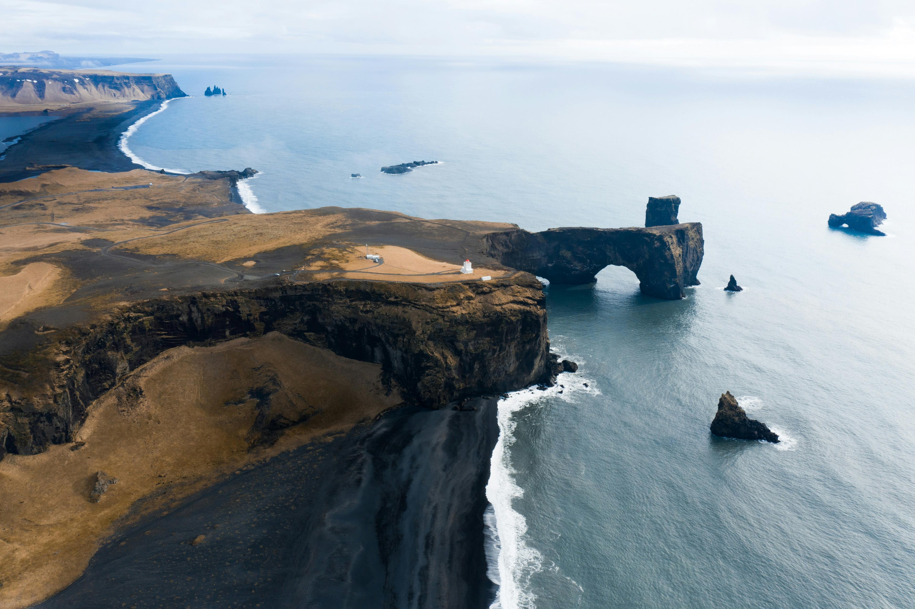 Aerial view of a rugged coastline with a distinctive arch rock formation in the sea, black sand beach, and a small lighthouse on the cliff.