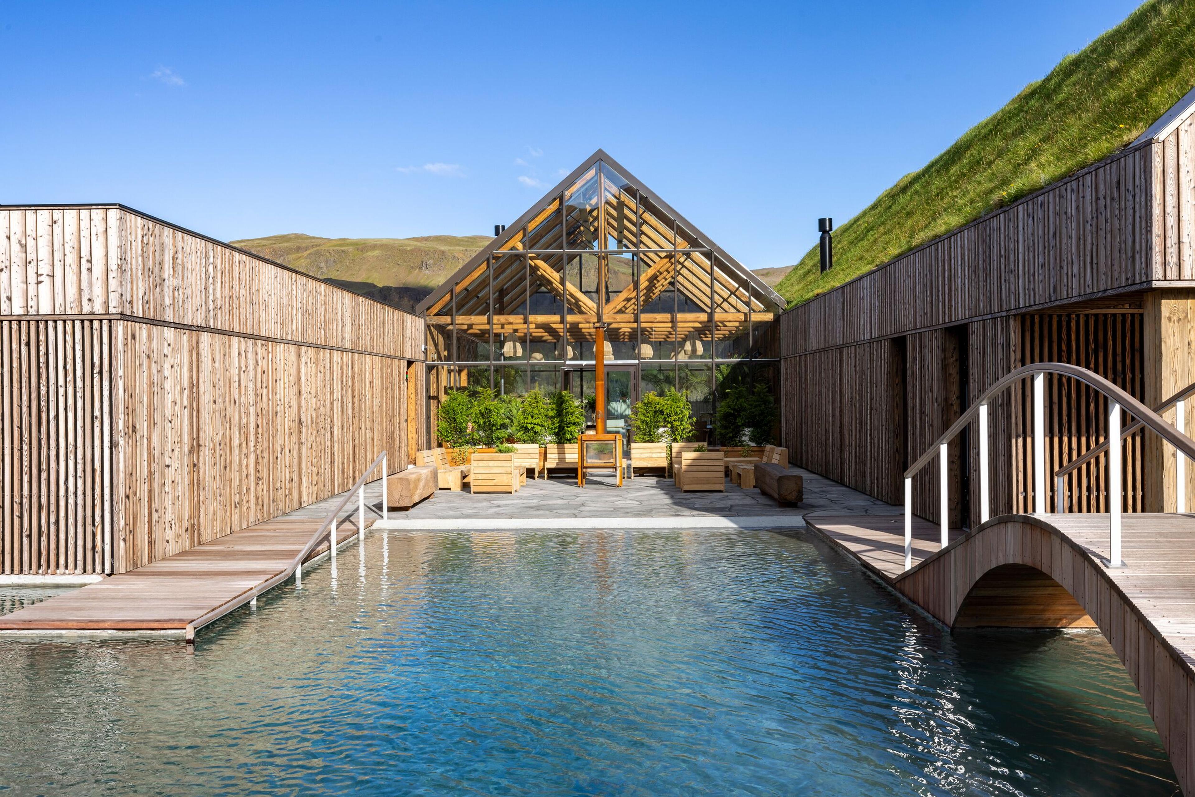 An outdoor swimming pool flanked by wooden buildings, featuring a central glass greenhouse, a grass-covered roof, and a small bridge.