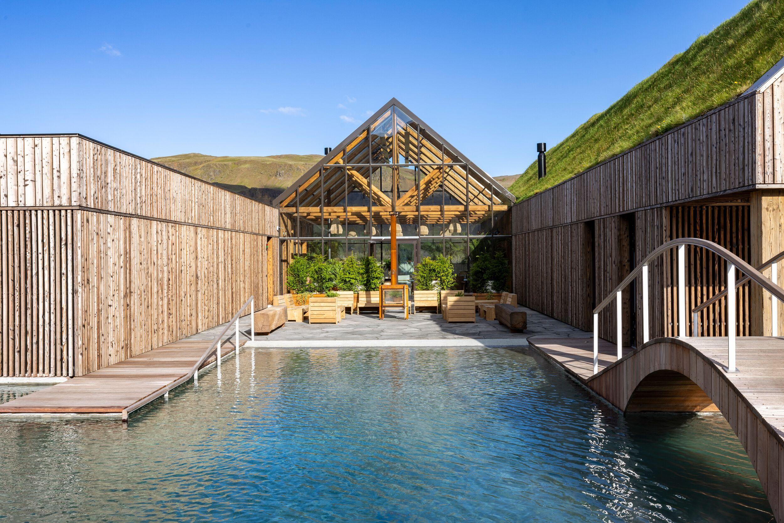 Outdoor pool with modern wooden buildings, one featuring a green roof and another a glass greenhouse, set against green hills.