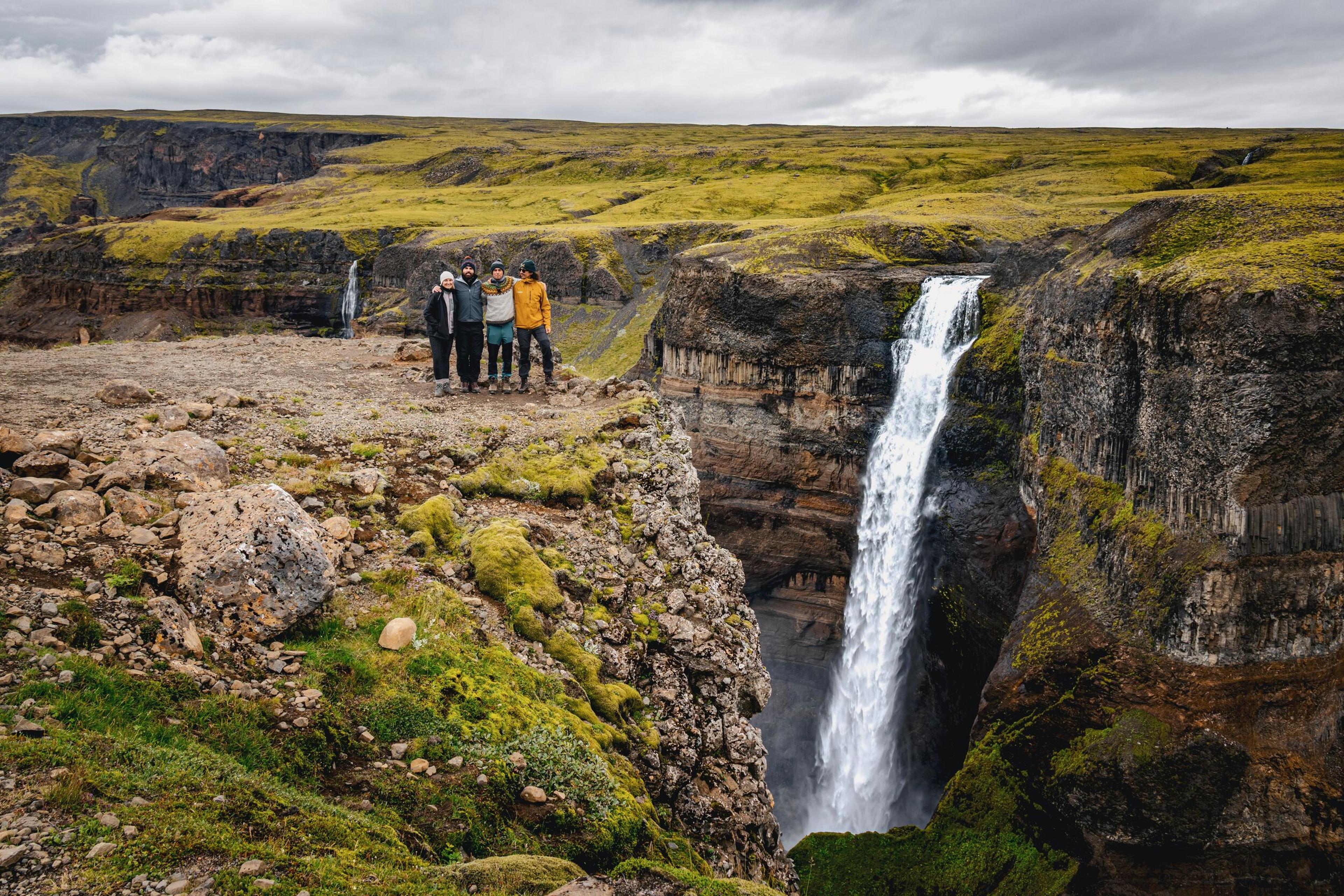 Five people stand on a rocky cliff overlooking a large waterfall in a green, mossy canyon.