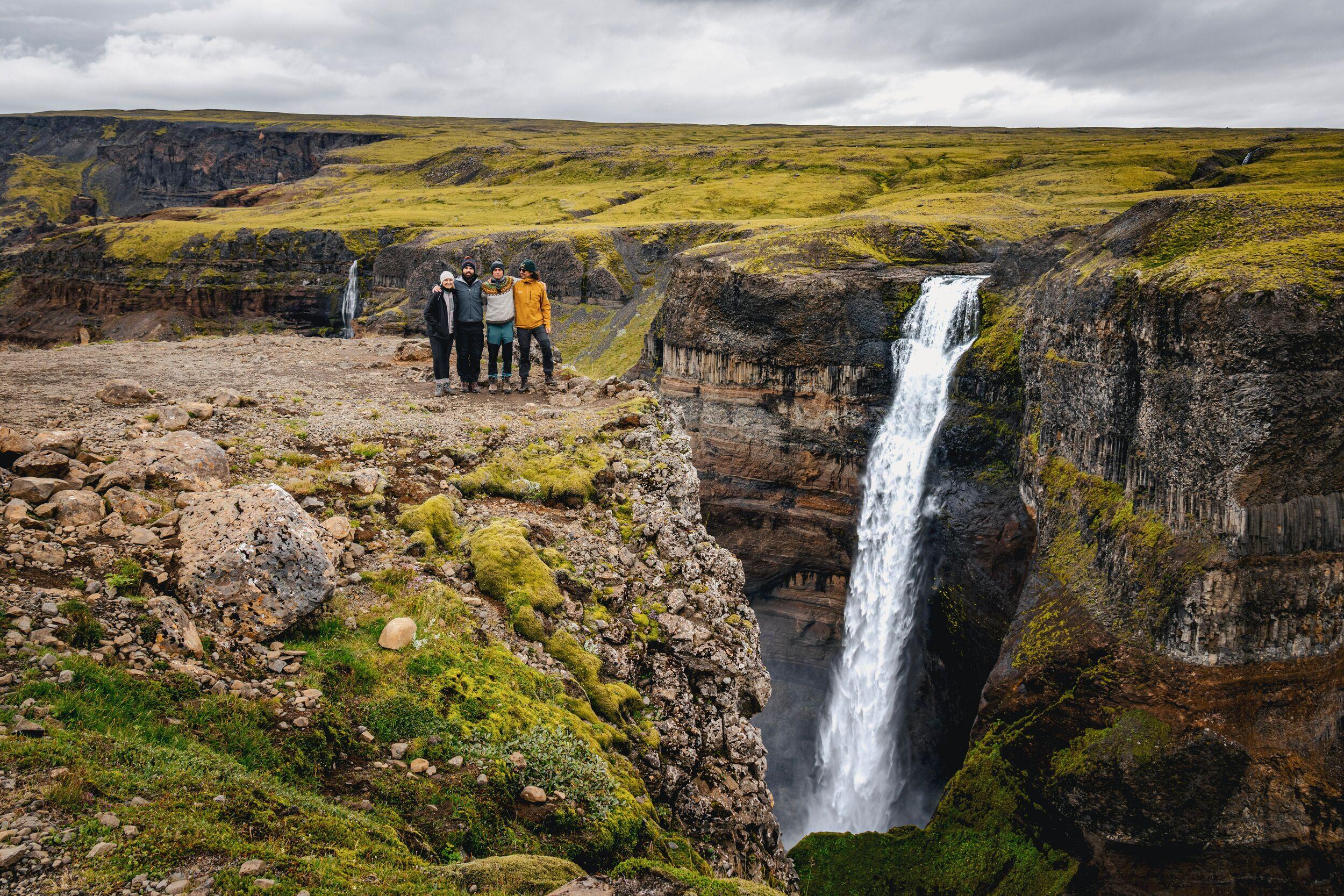 Five people stand on a rocky cliff overlooking a large waterfall in a green, mossy canyon.