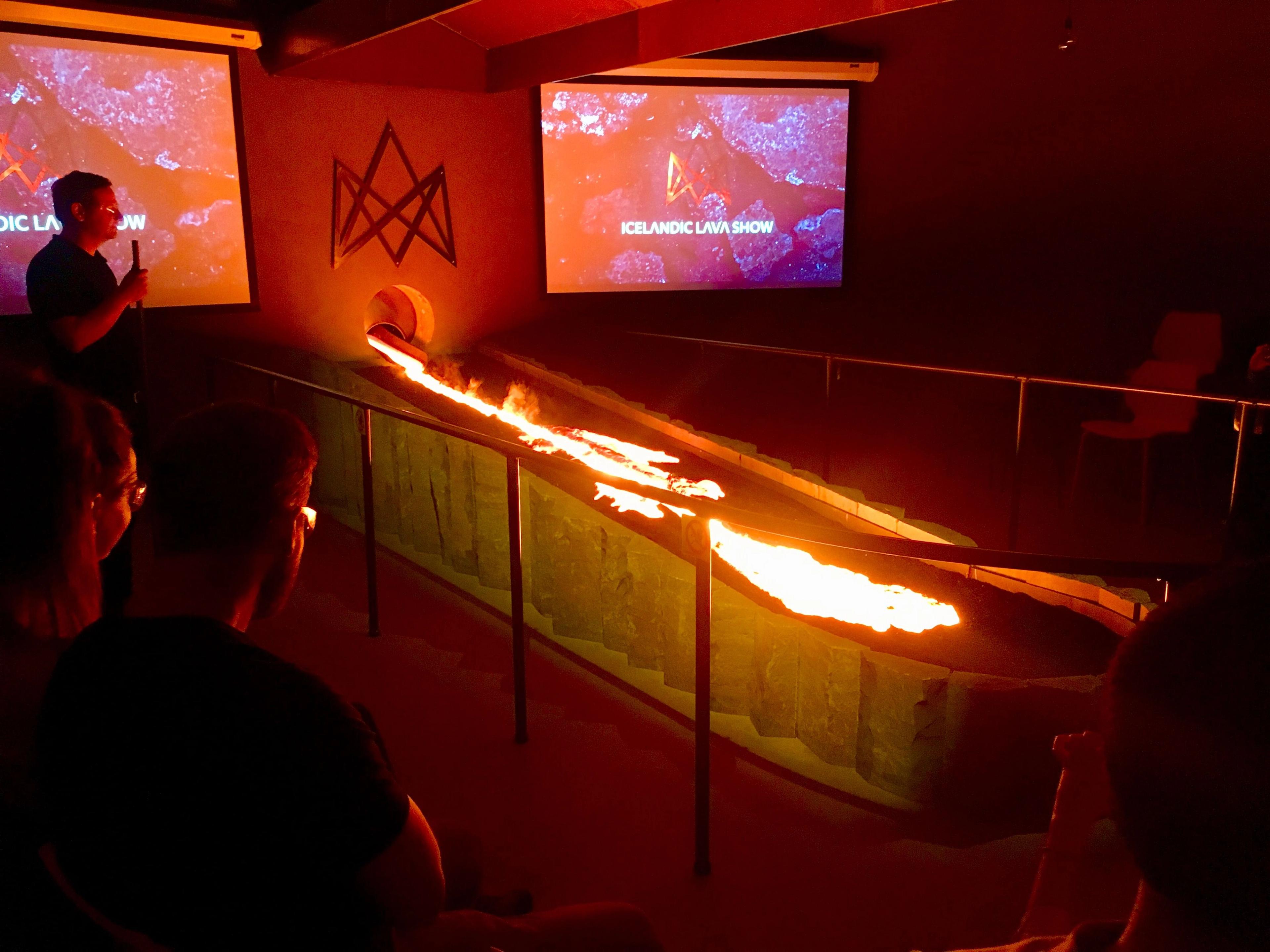Attendees watch as glowing molten lava pours from a wall and flows down a channel at the Icelandic Lava Show in a dimly lit room.