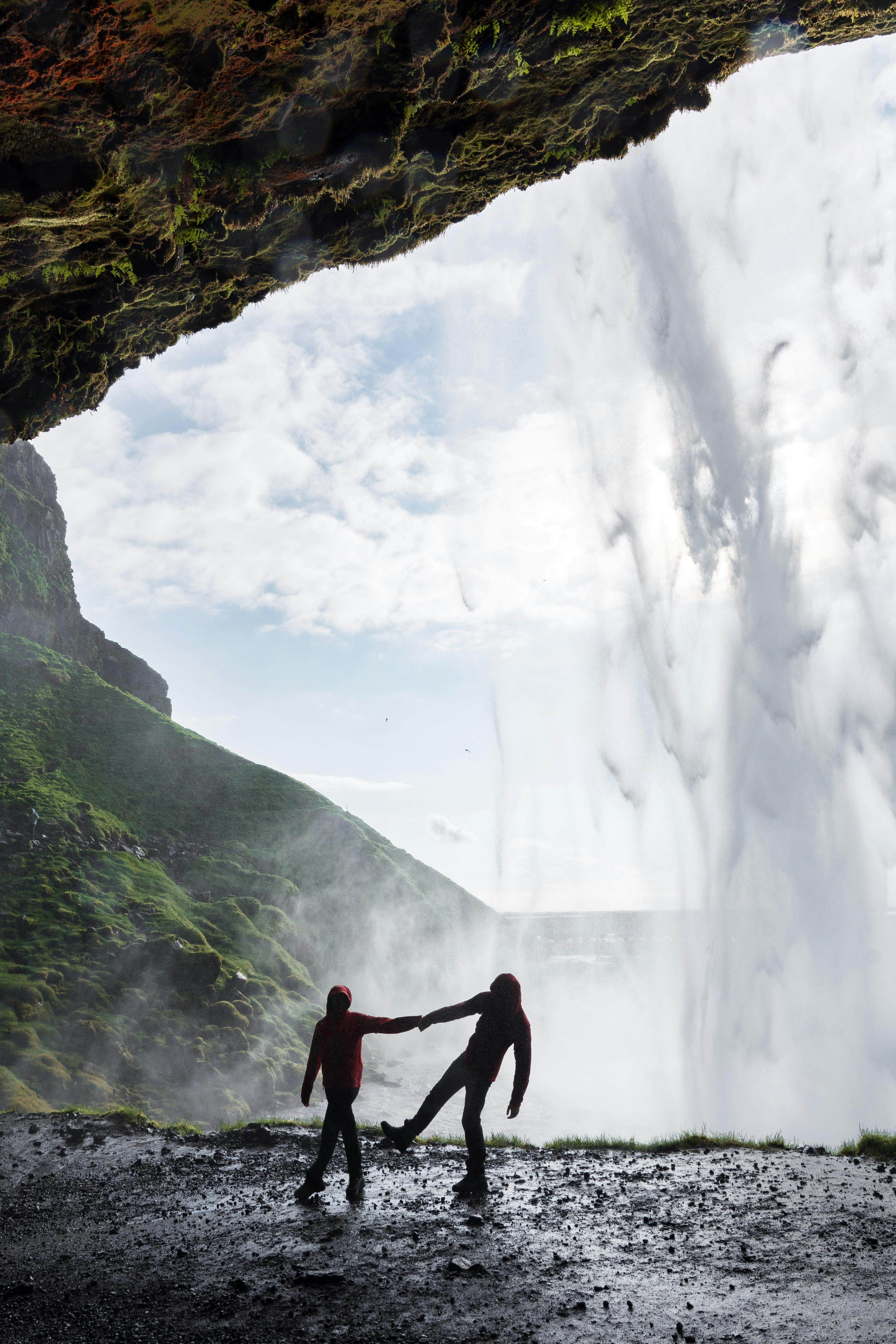 Two people silhouetted behind Seljalandsfoss waterfall in Iceland, standing on a wet, rocky surface with mist in the air, creating a dramatic and adventurous scene.