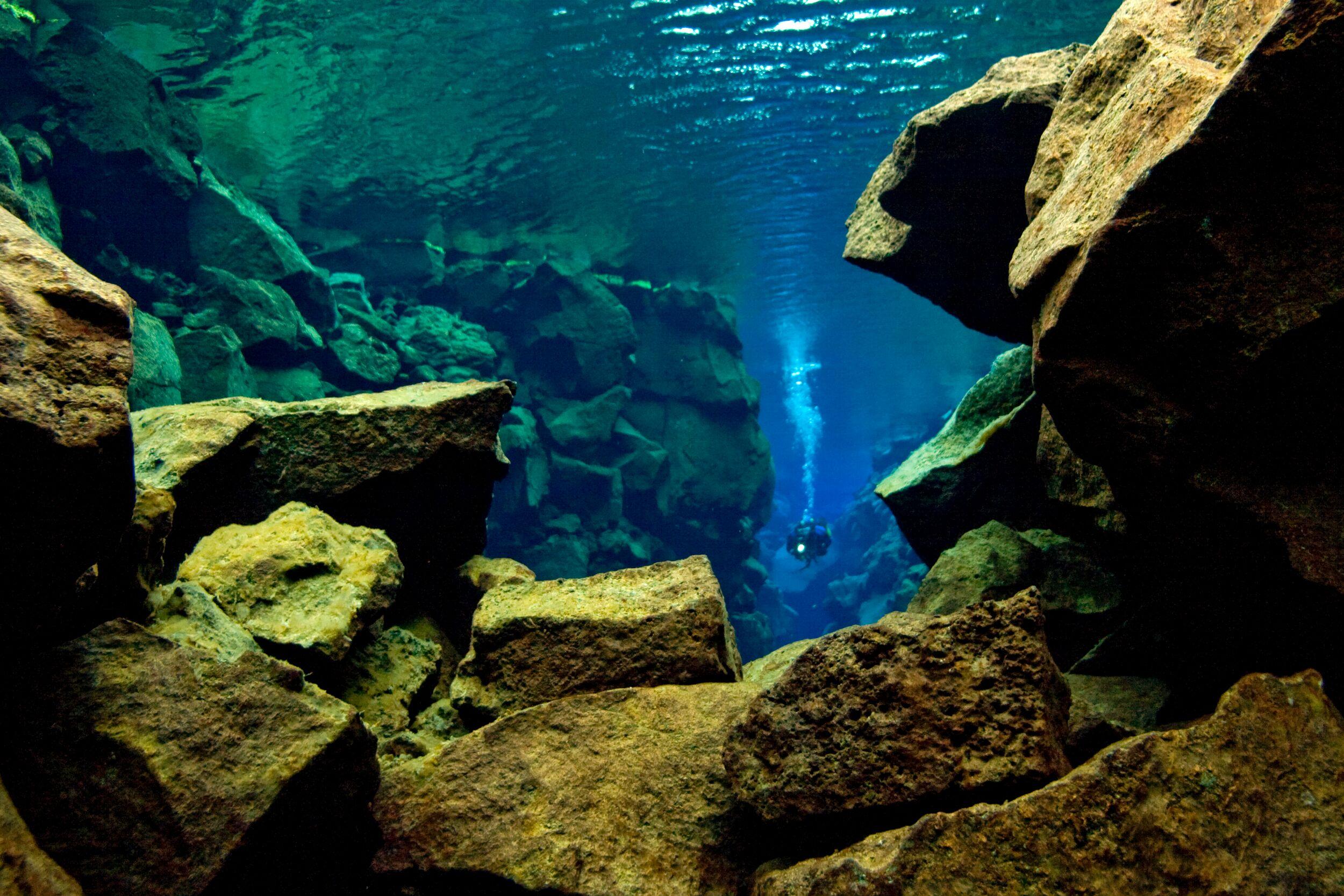 A diver in clear blue-green water ascends through a rocky underwater fissure.