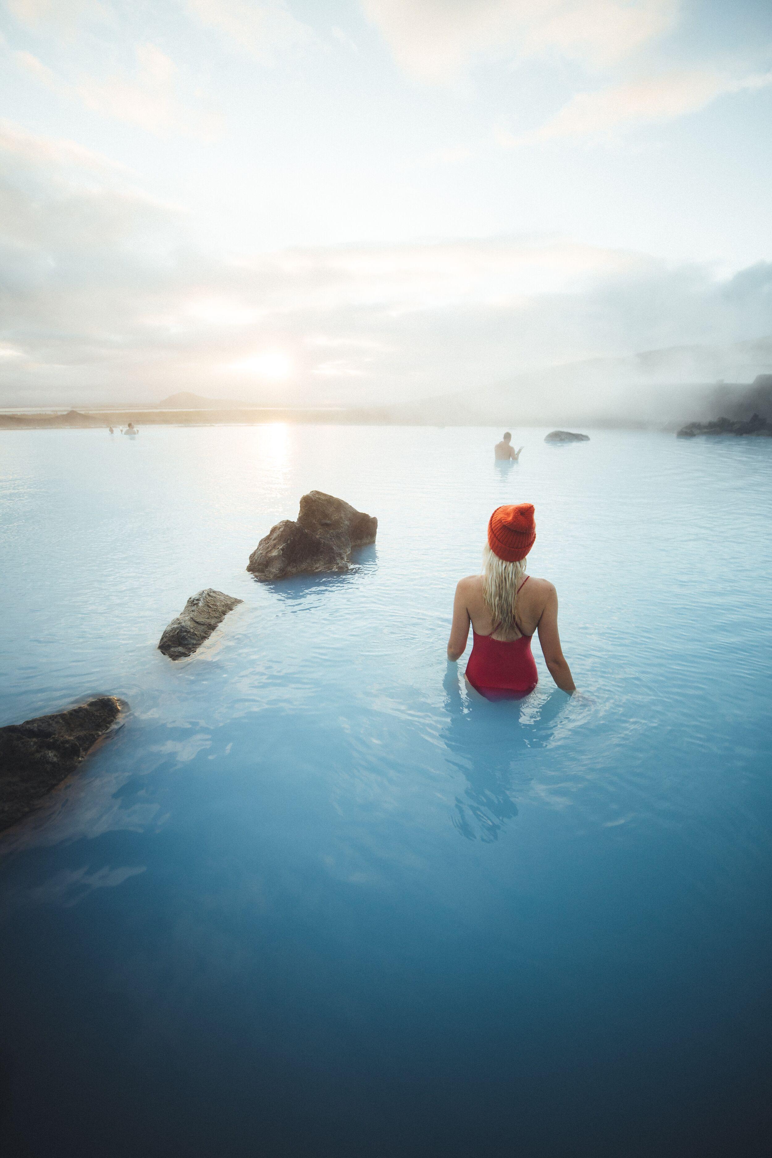 Woman in red swimsuit and orange beanie wades in a steamy, vibrant blue geothermal pool, with rocks and distant figures under a bright sky.
