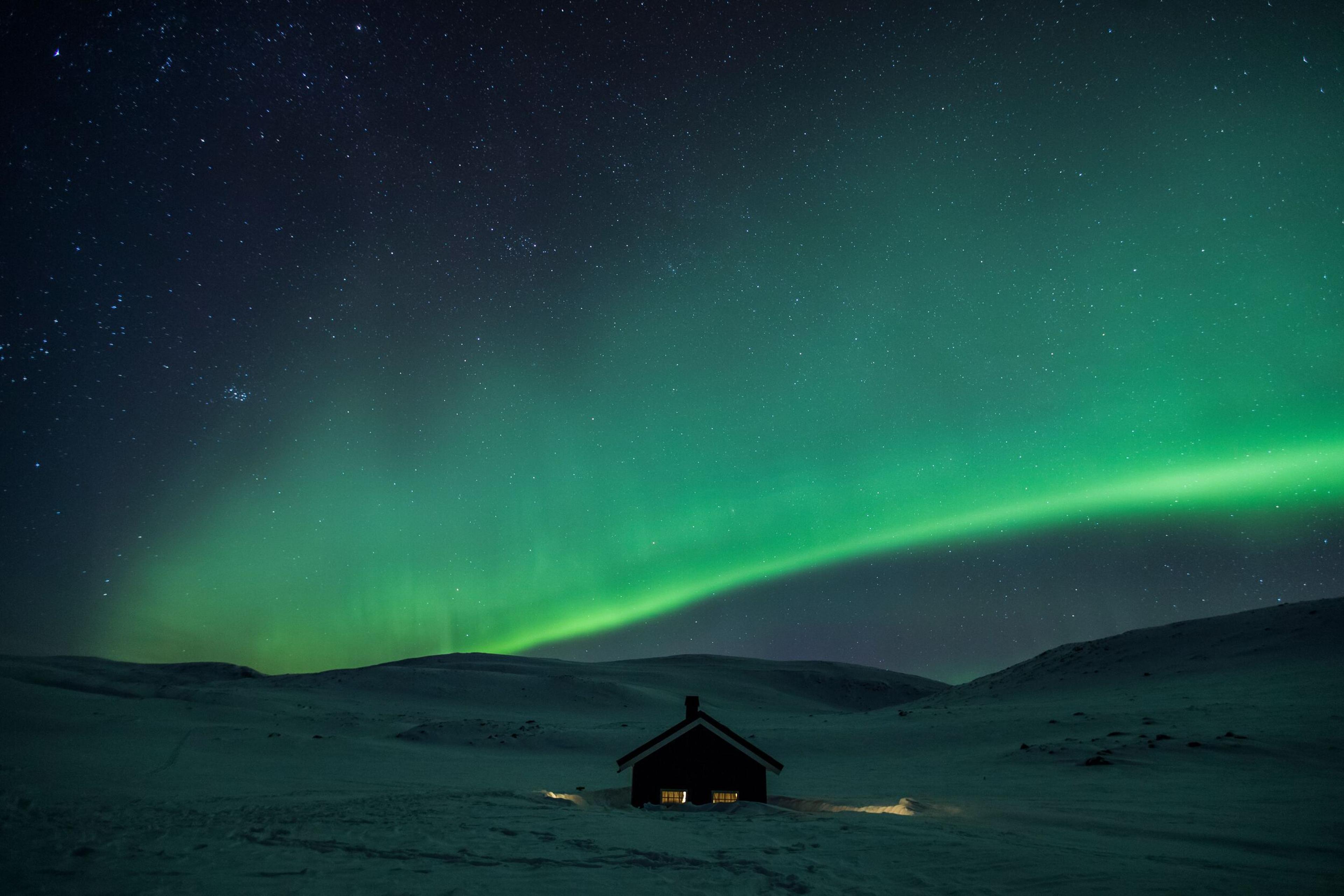 A small house in a snowy landscape under a vibrant green aurora borealis and a starry night sky.