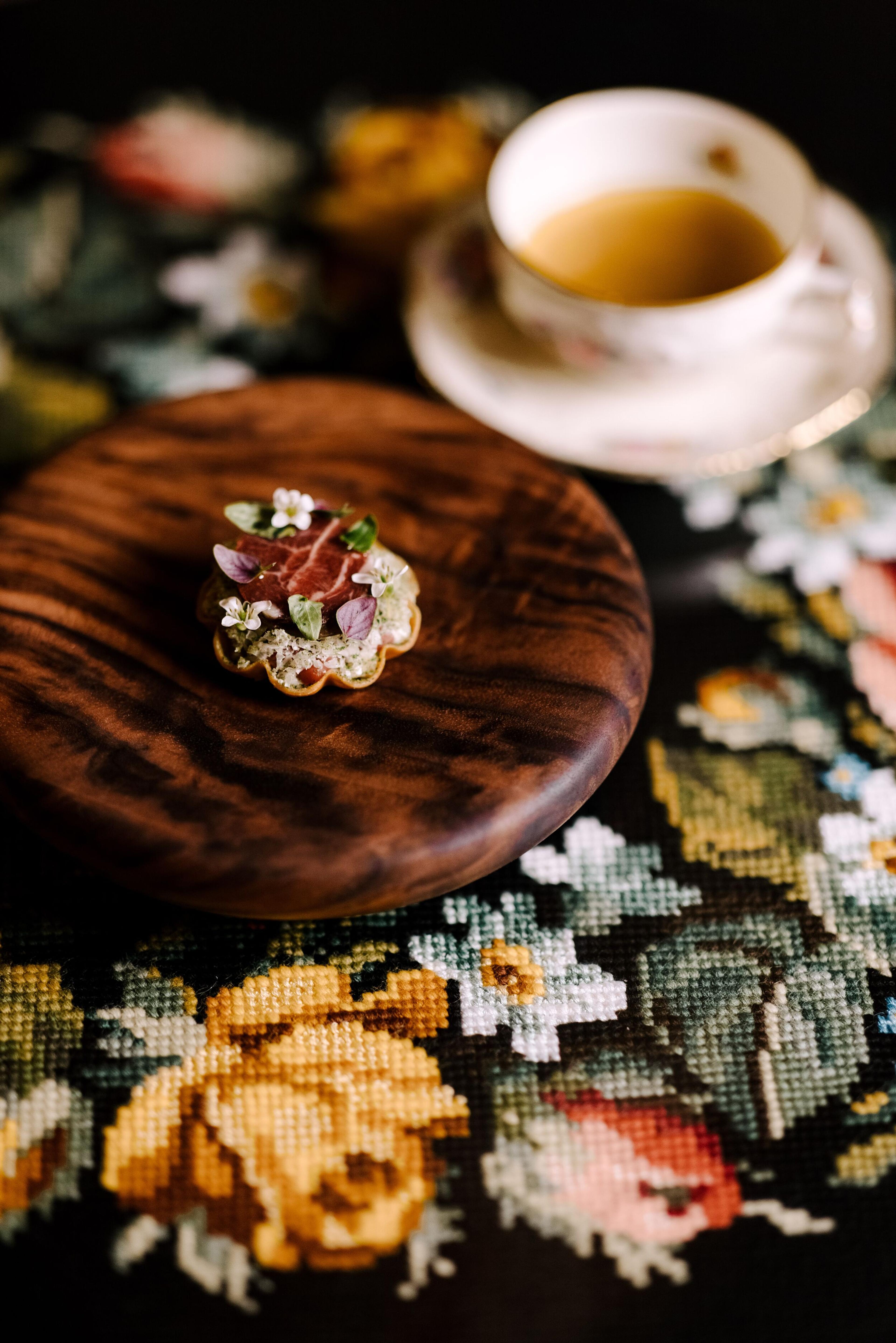 A gourmet tartlet with cured meat, herbs, and flowers on a wooden plate, alongside a teacup on a floral embroidered tablecloth.
