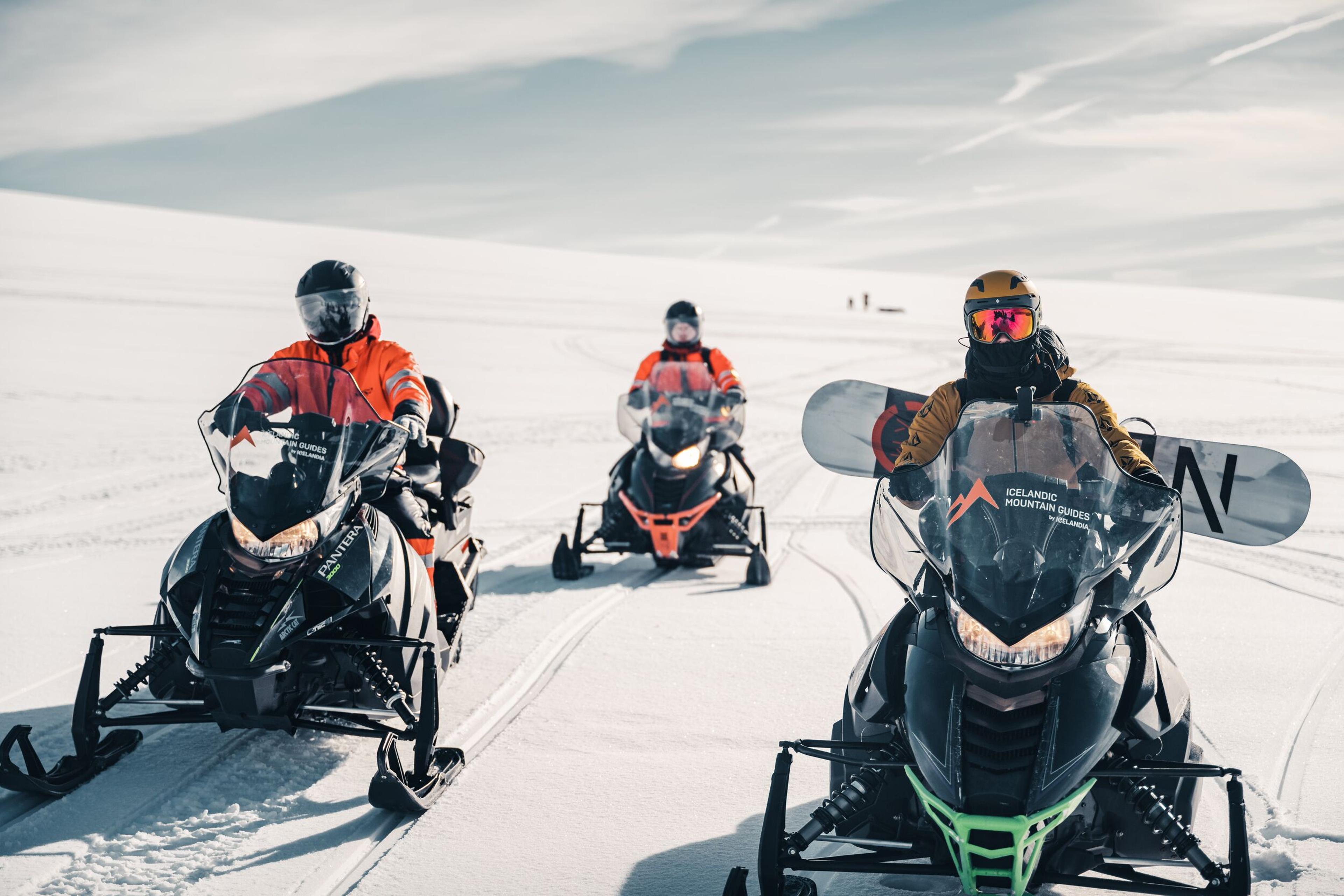 Three people on snowmobiles in Iceland, one carrying a snowboard, ready for an arctic adventure.