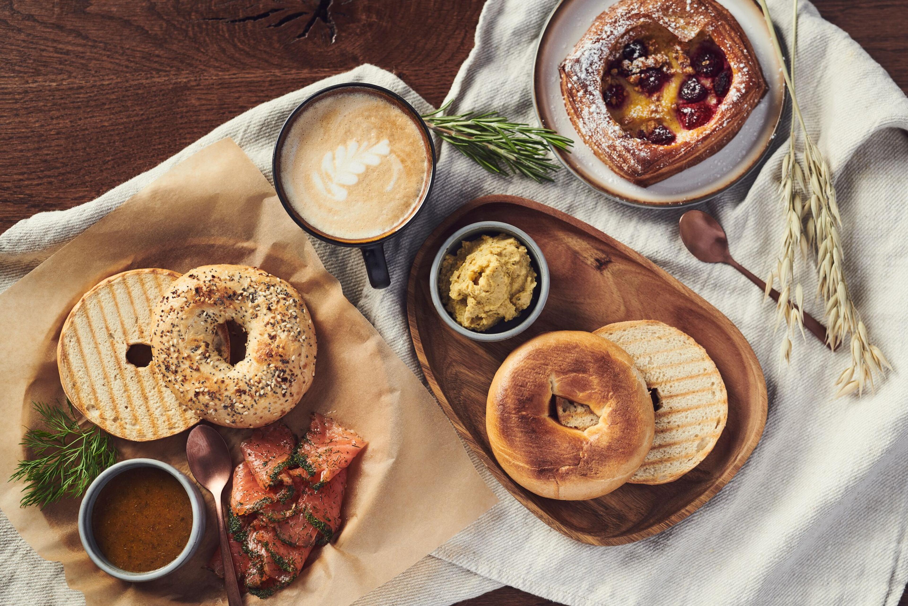 A cozy and appetizing spread of Nordic cuisine, featuring grilled bread, a bagel, smoked salmon, a bowl of soup, spreads, and a sweet pastry, paired with a latte, presented on wooden boards and a rustic table, evoking a culinary experience at Sky Lagoon Iceland.