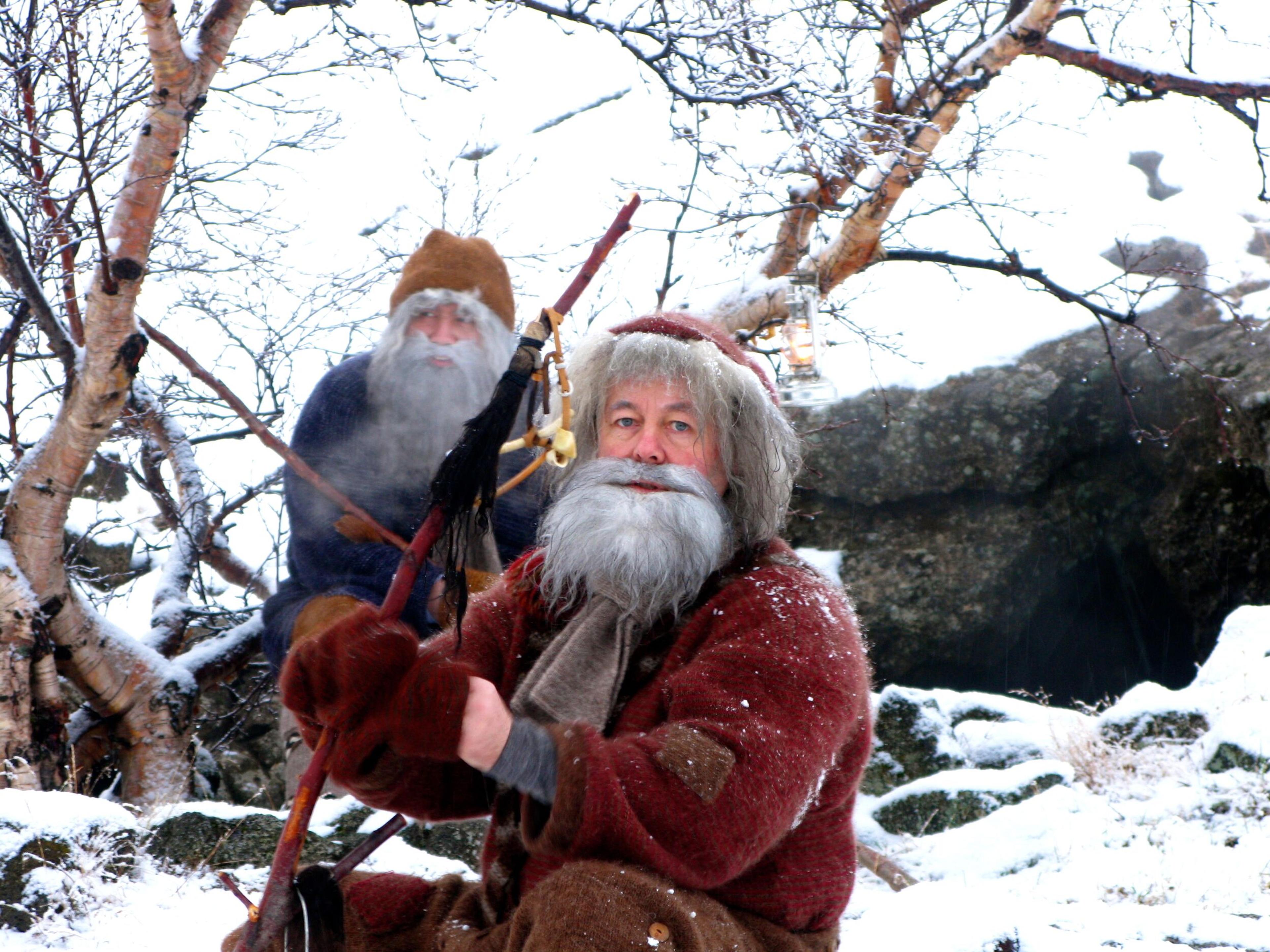"Two figures dressed in traditional Icelandic Yule Lad costumes, with long white beards and colorful robes, standing in a snowy landscape, one in the foreground holding a wooden staff, and the other in the background amidst bare winter branches.