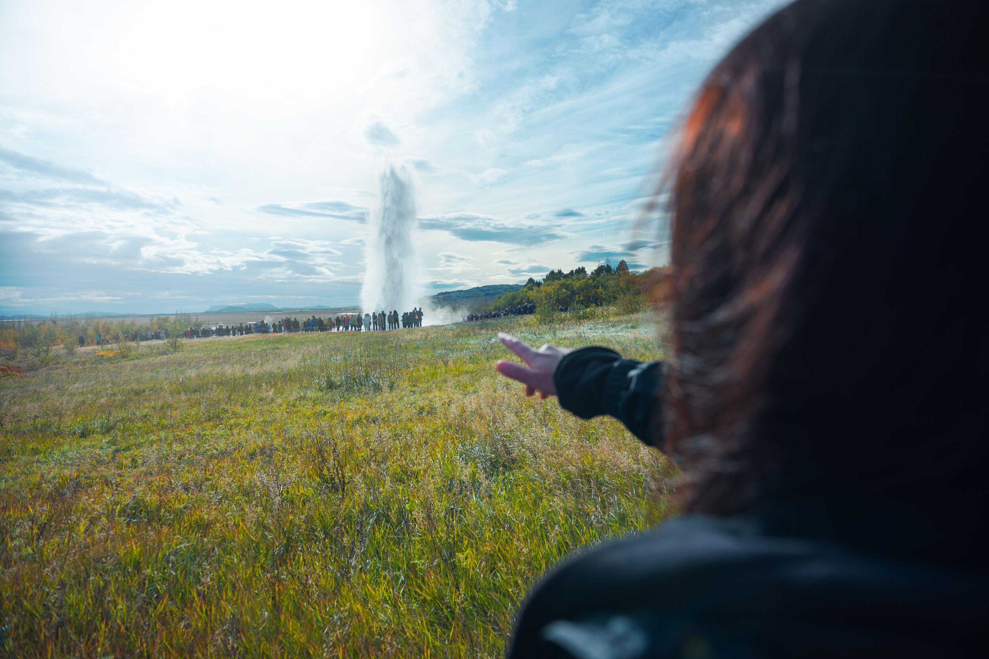 A person is gesturing towards a distant geyser erupting in a grassy field under a blue sky with clouds.