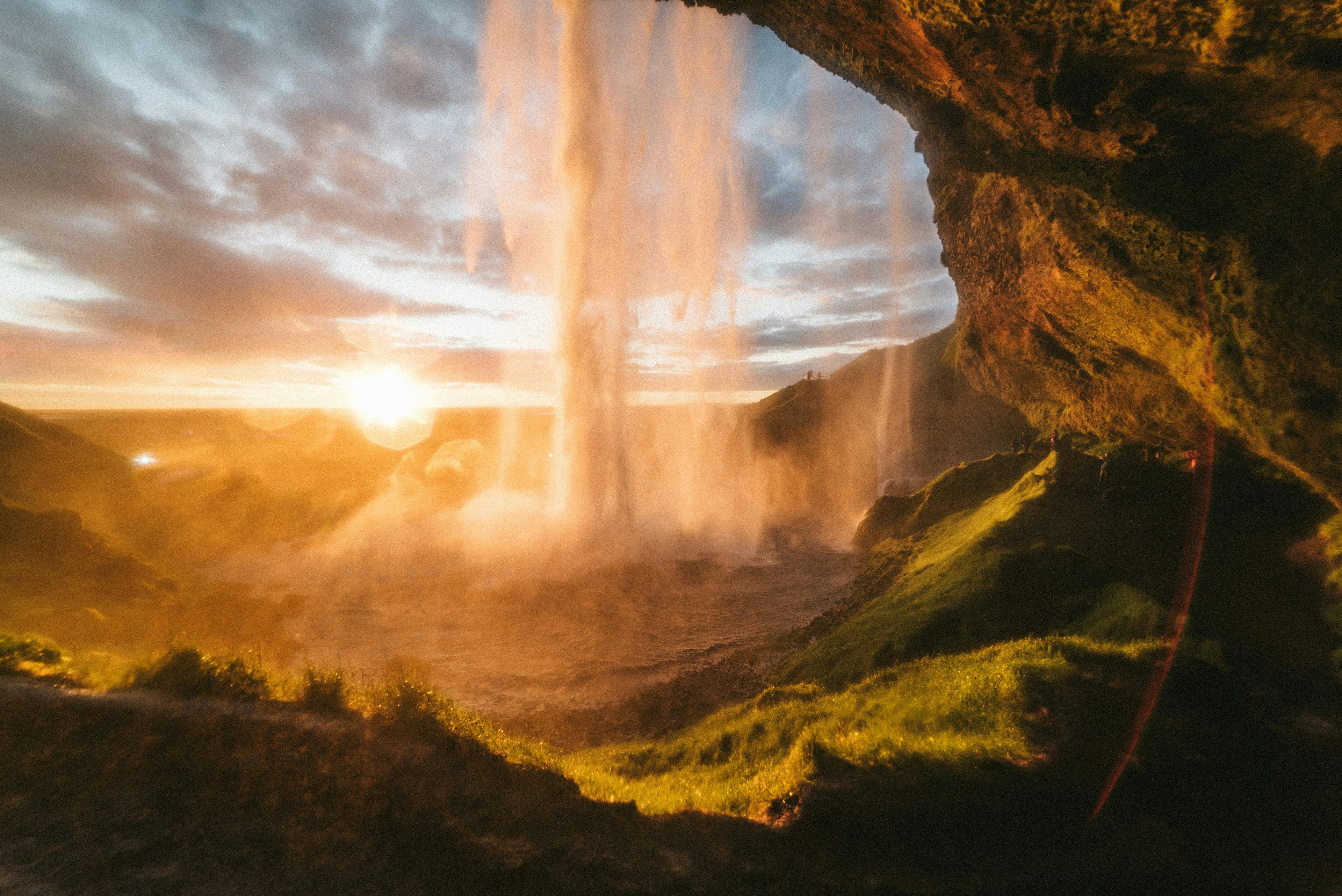 View of Seljalandsfoss waterfall from behind the cascade at sunset, with golden light illuminating the mist and green landscape.