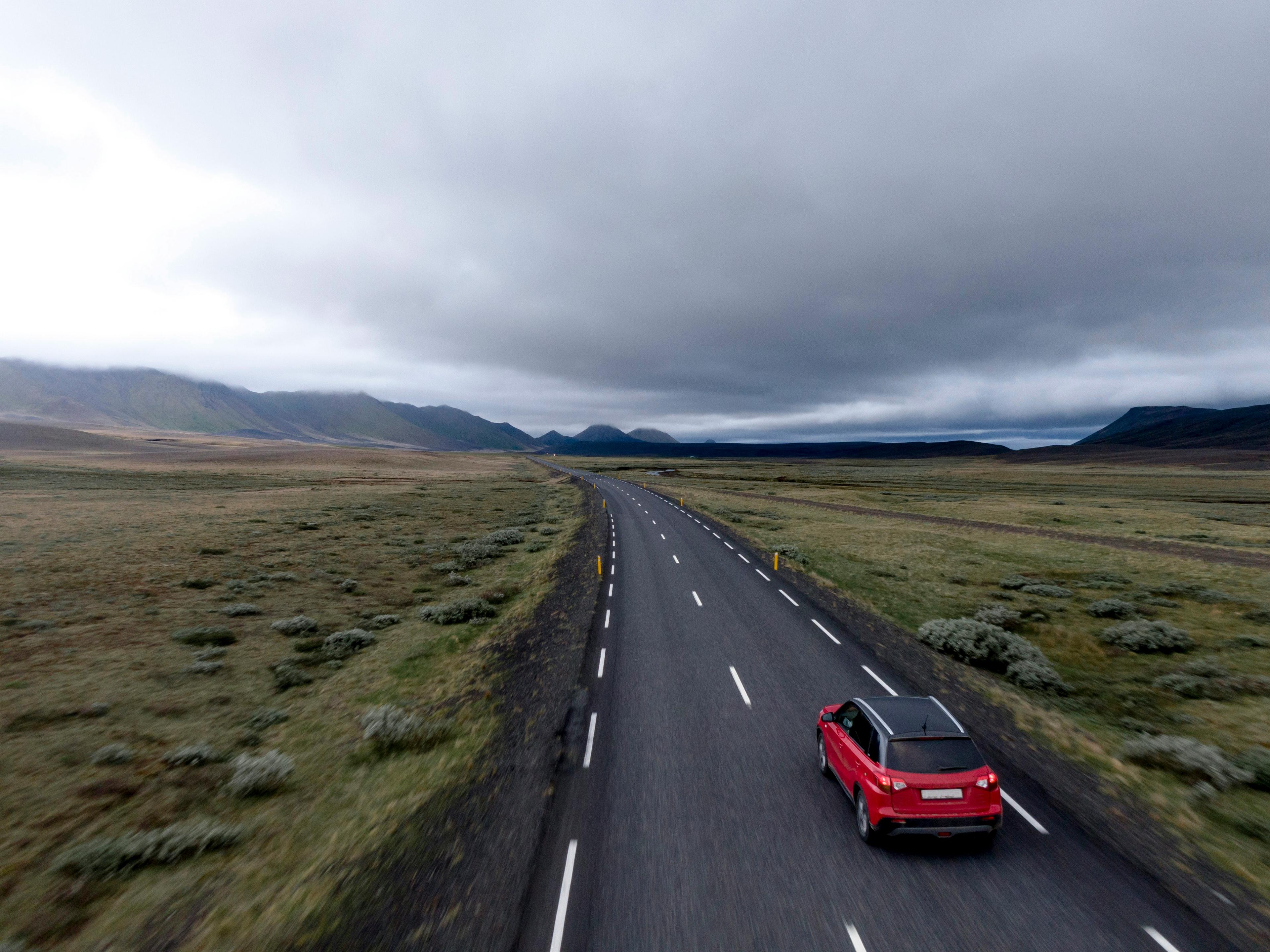 A red SUV driving along a long, empty road that stretches through vast, open plains with rugged green landscapes on either side. 