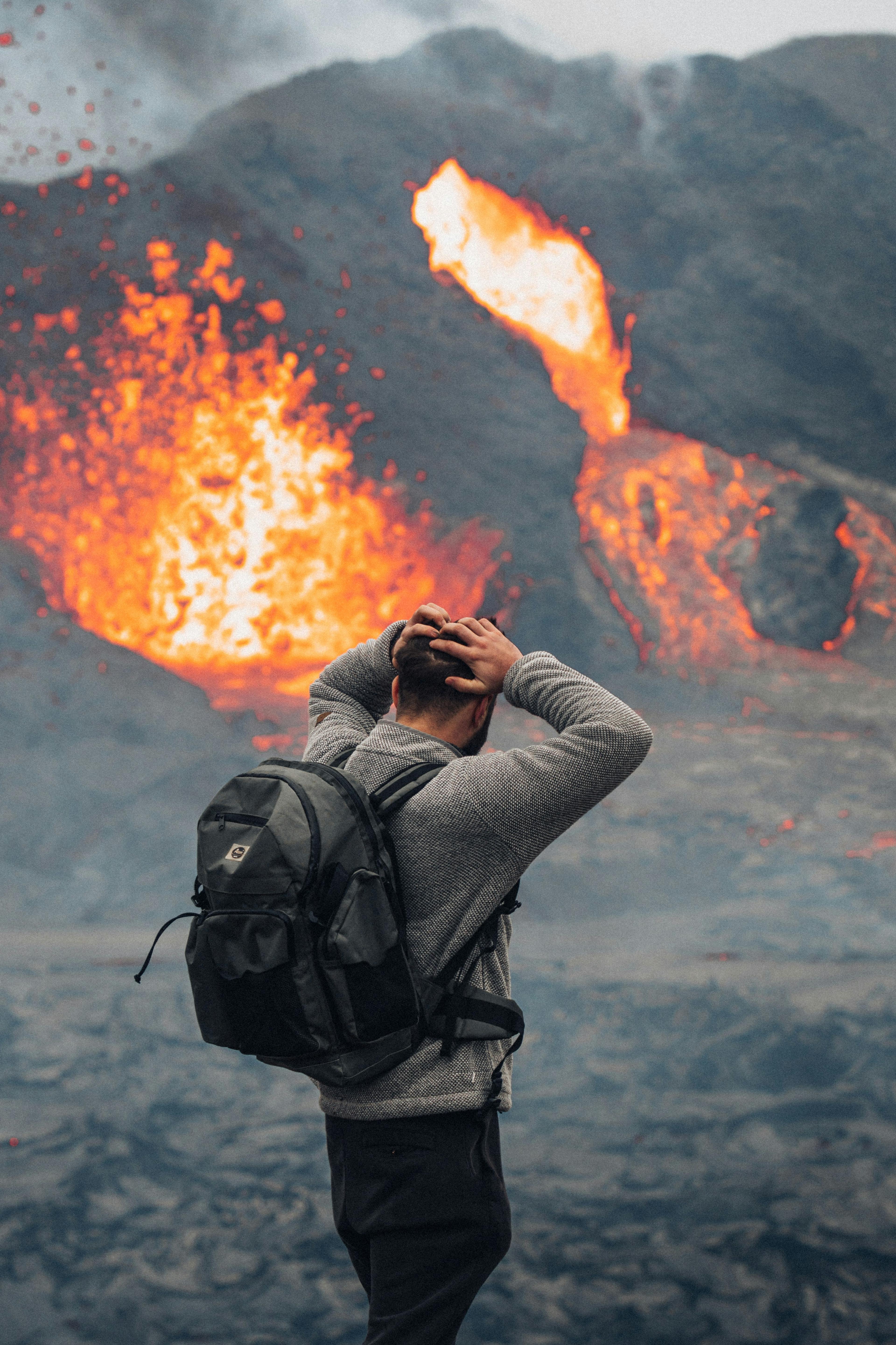 An awestruck spectator with a backpack grasps his head in disbelief at the intense and magnificent eruption before him, as Fagradalsfjall spews fiery lava high into the air.