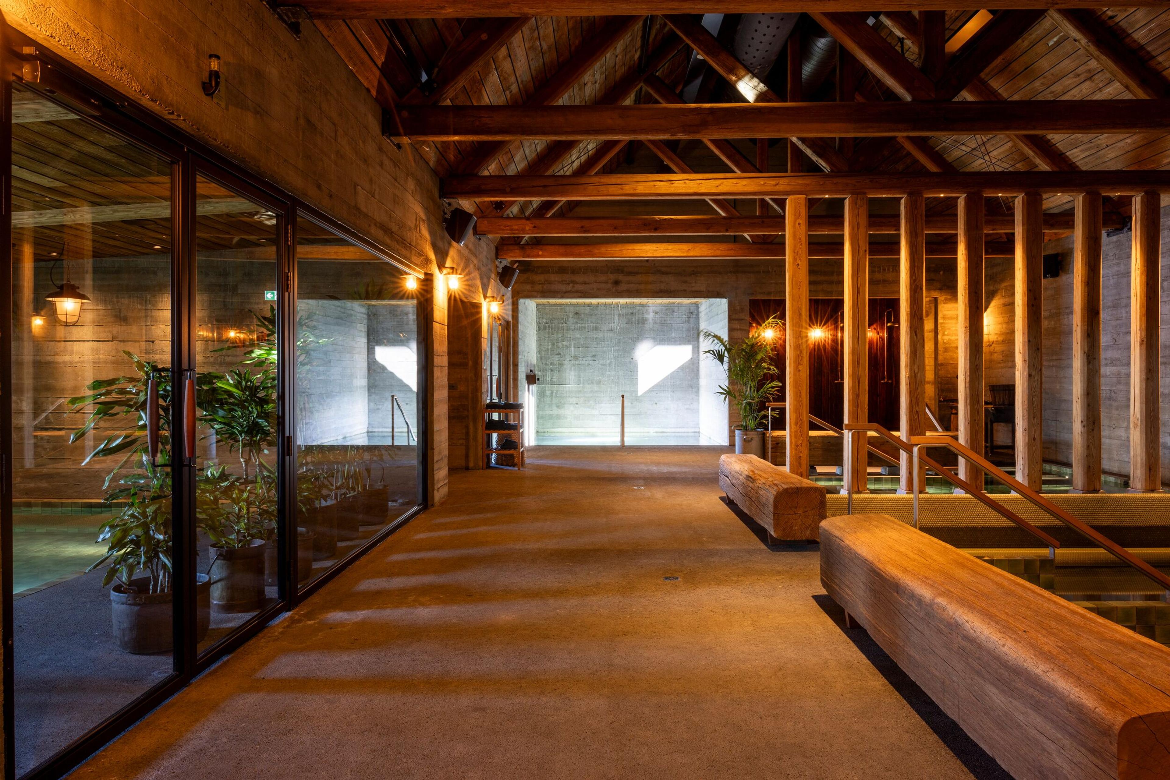 Warmly lit modern spa interior with concrete walls, exposed wood beams, a glass-enclosed pool, and wooden benches by sunken water features.