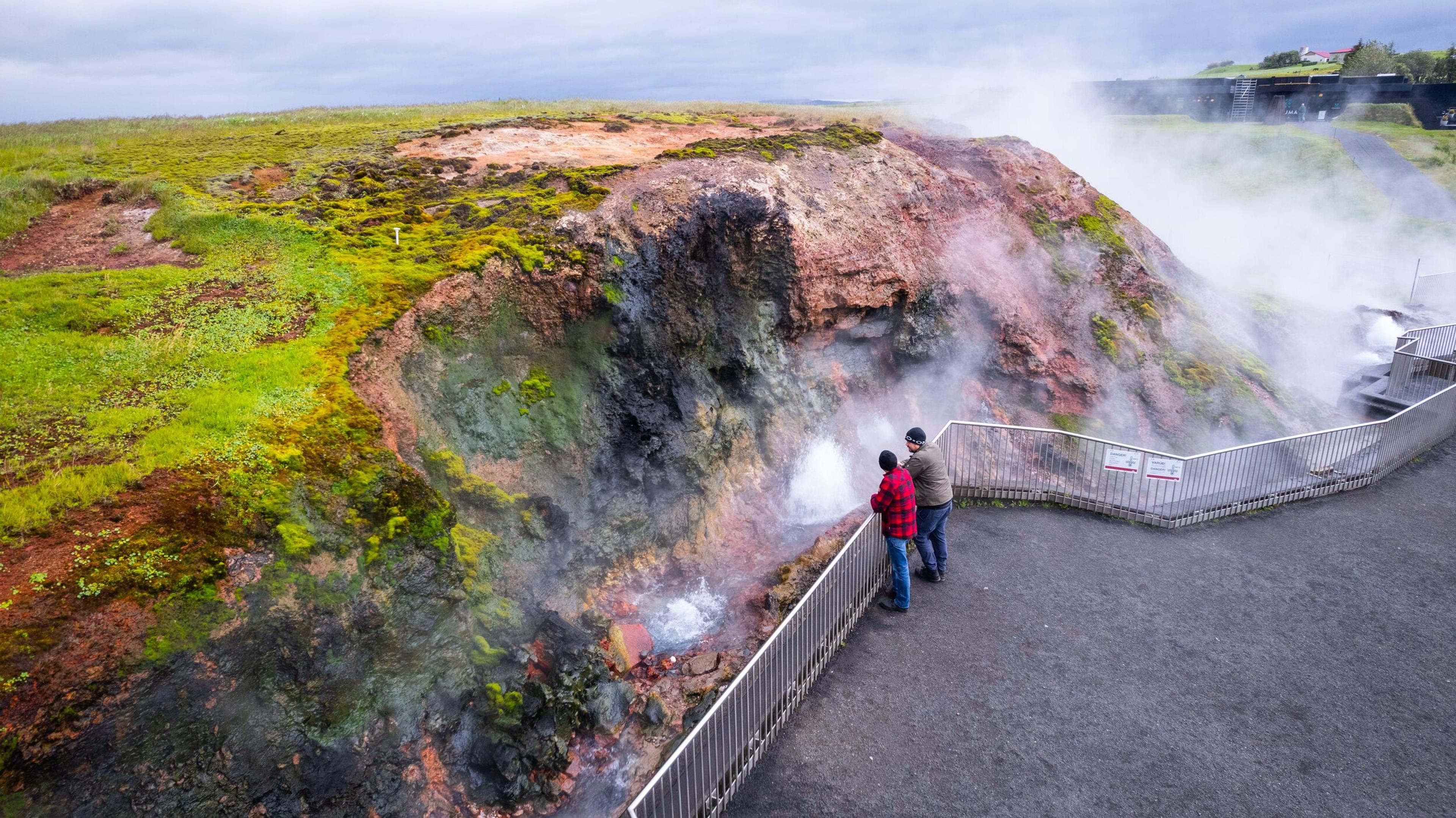 Visitors observing Deildartunguhver geothermal hot spring on the Silver Circle, featuring steaming vents surrounded by moss-covered rocks and vibrant geothermal colors, with visitors observing from a safe fenced platform.