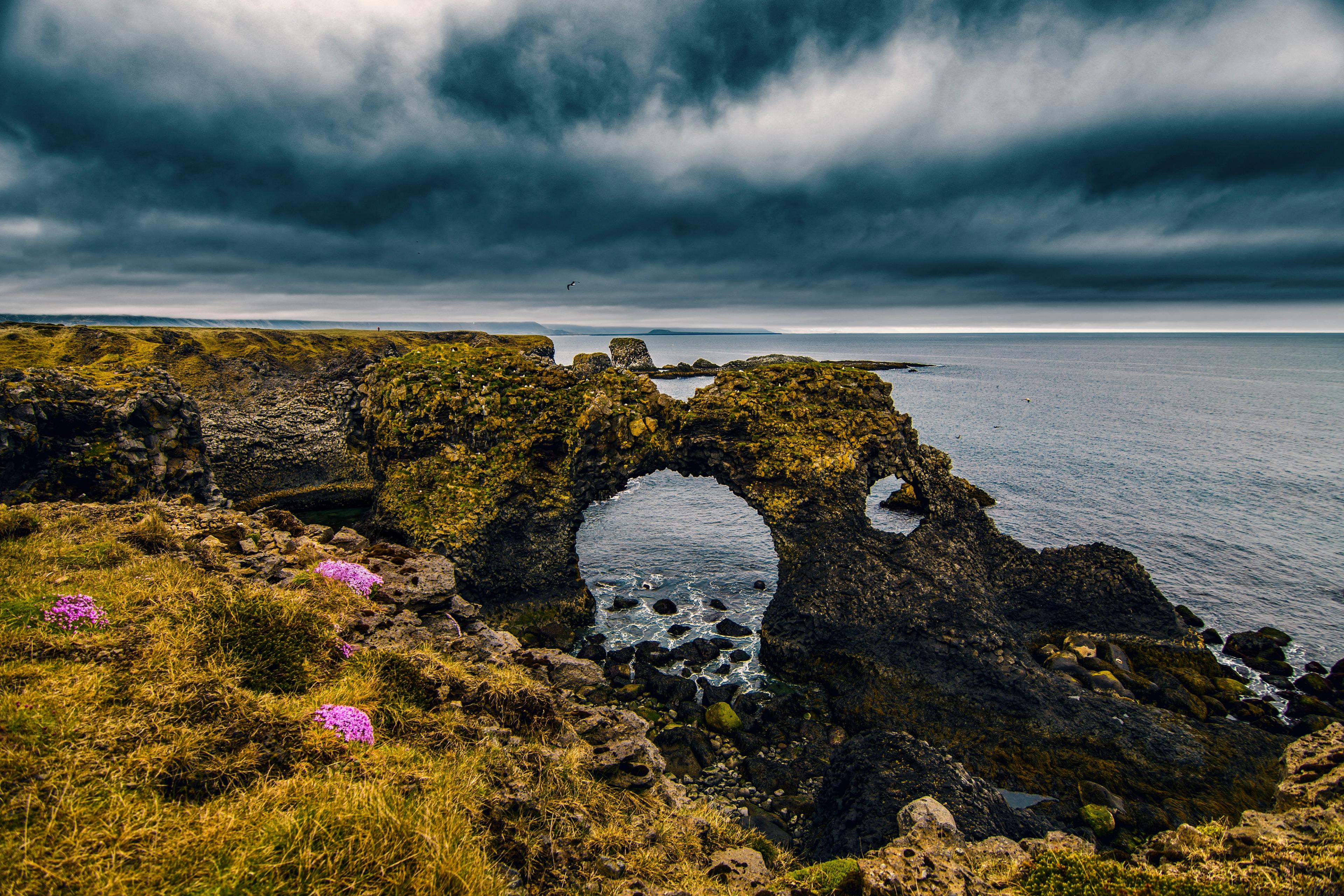 A moss-covered natural rock arch on a rugged coastline under a dramatic, dark cloudy sky, with the ocean visible beyond.