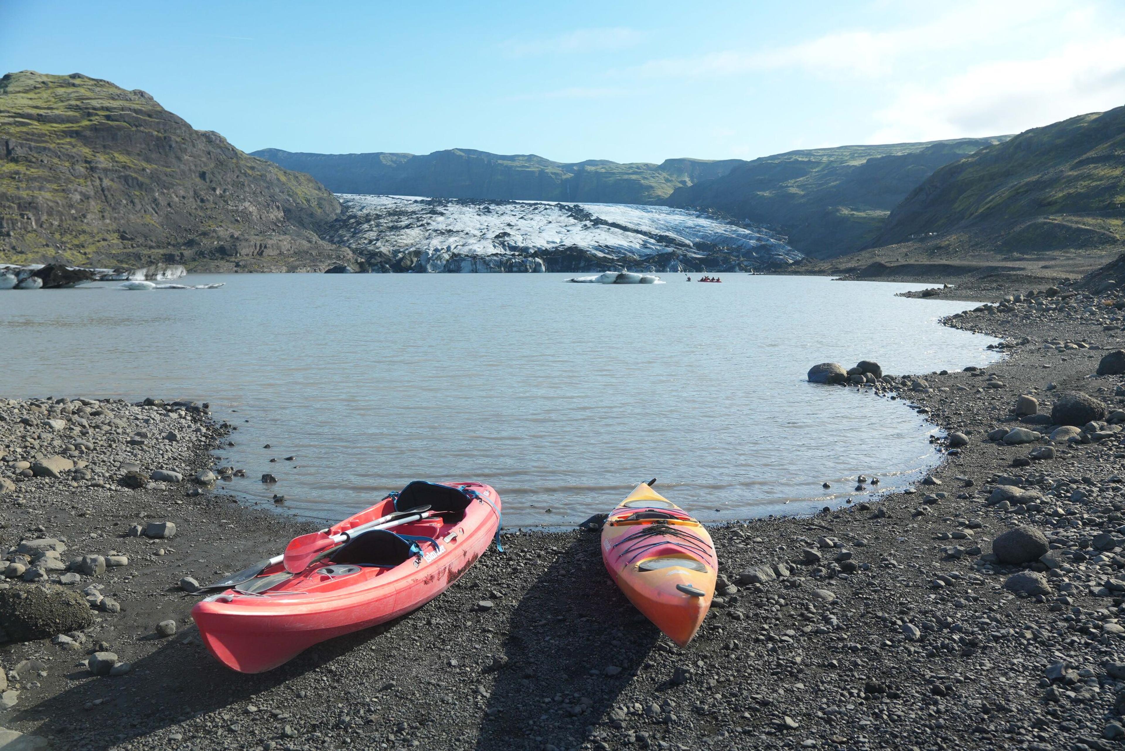 Two kayaks on a rocky shore of a glacial lake with a large glacier and mountains in the background.