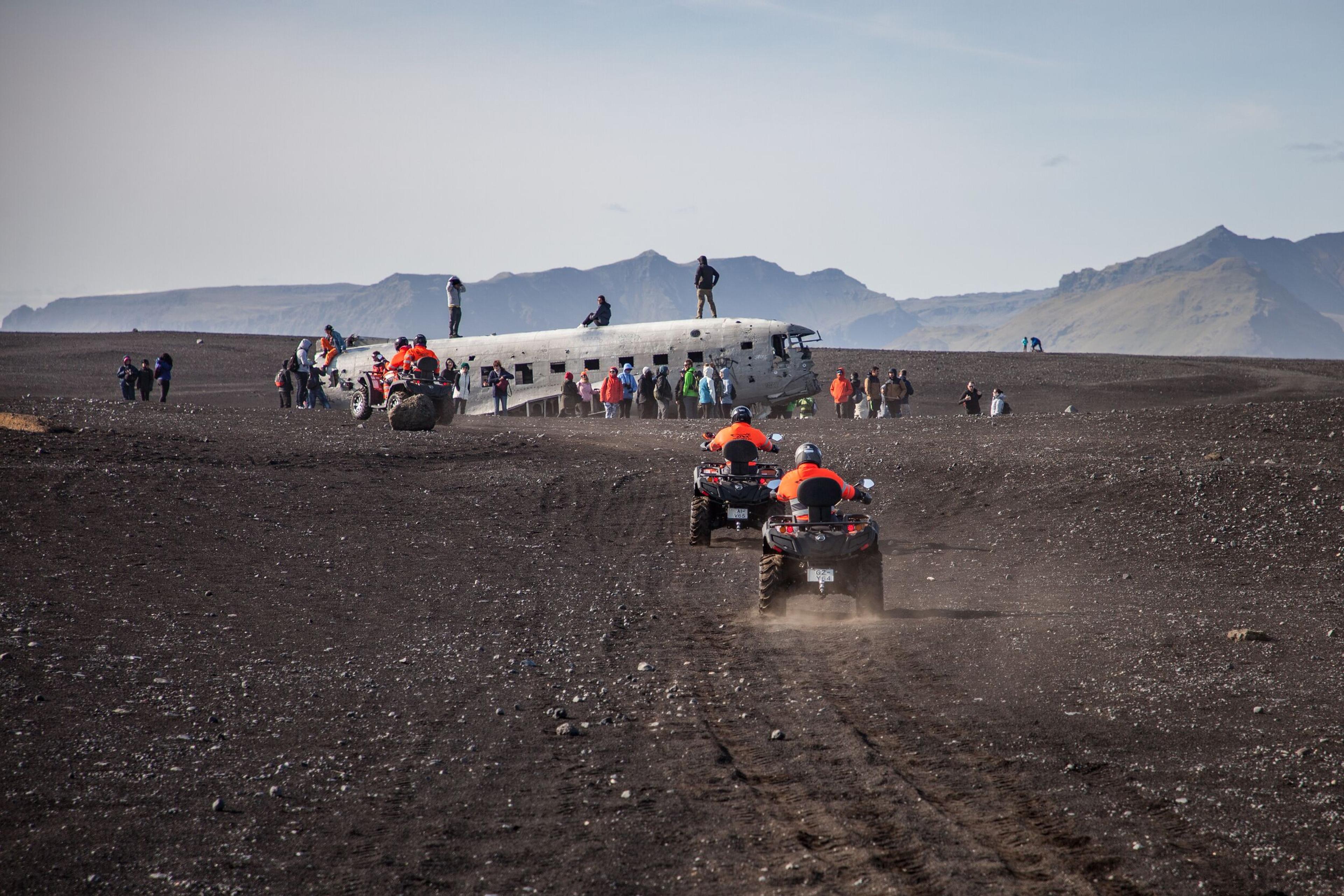 Two ATV riders in bright orange gear approach the famous Solheimasandur plane wreck on Iceland’s black sand desert, where a crowd of visitors explores the abandoned aircraft.