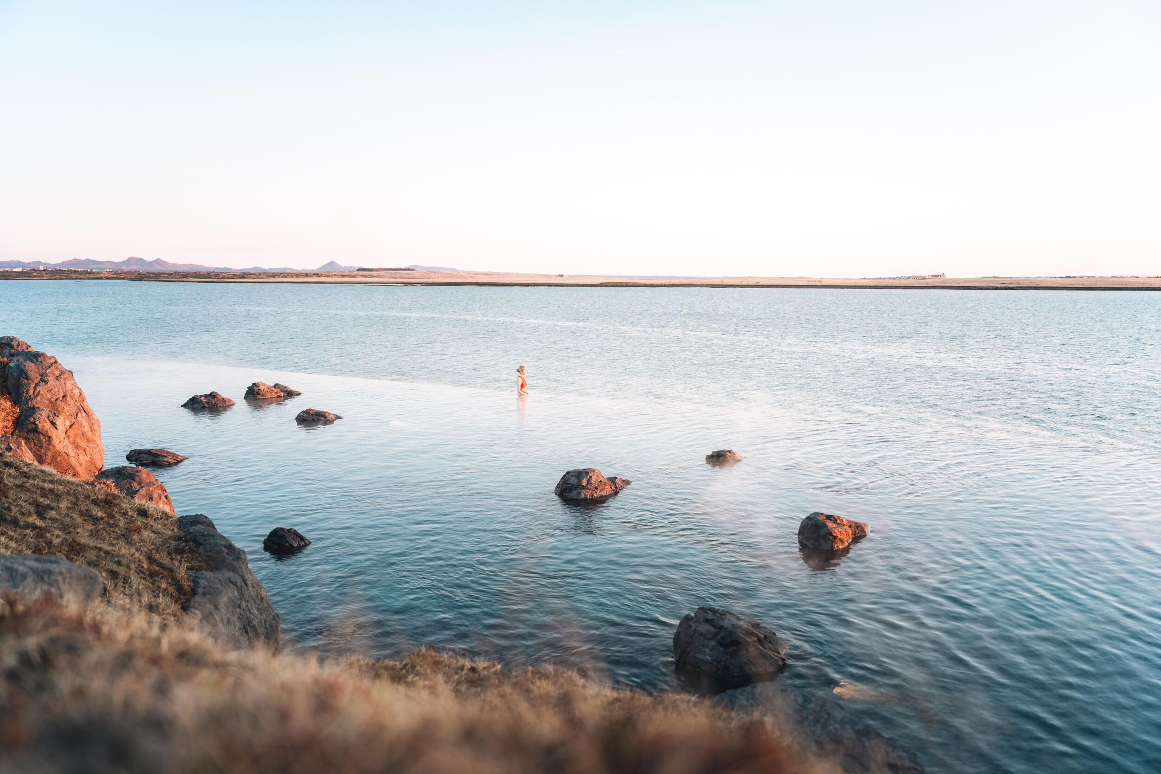 A solitary figure stands in the calm, expansive waters of Sky Lagoon, surrounded by dark rocks and the vast landscape of Iceland, under a pastel sky.