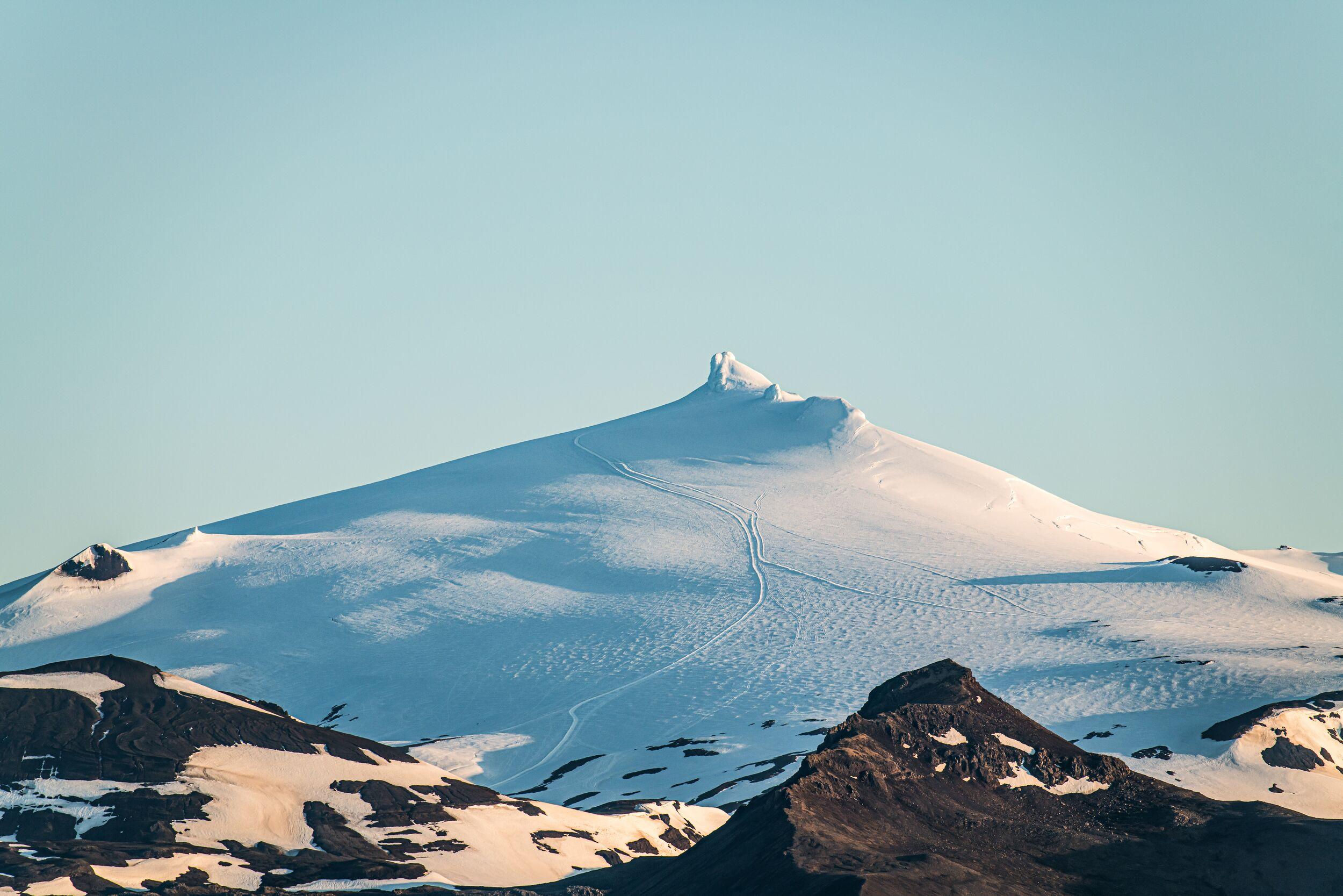 Snow-capped Snæfellsjökull glacier with a pointed peak and hiking tracks, above dark rocky hills under a clear sky.