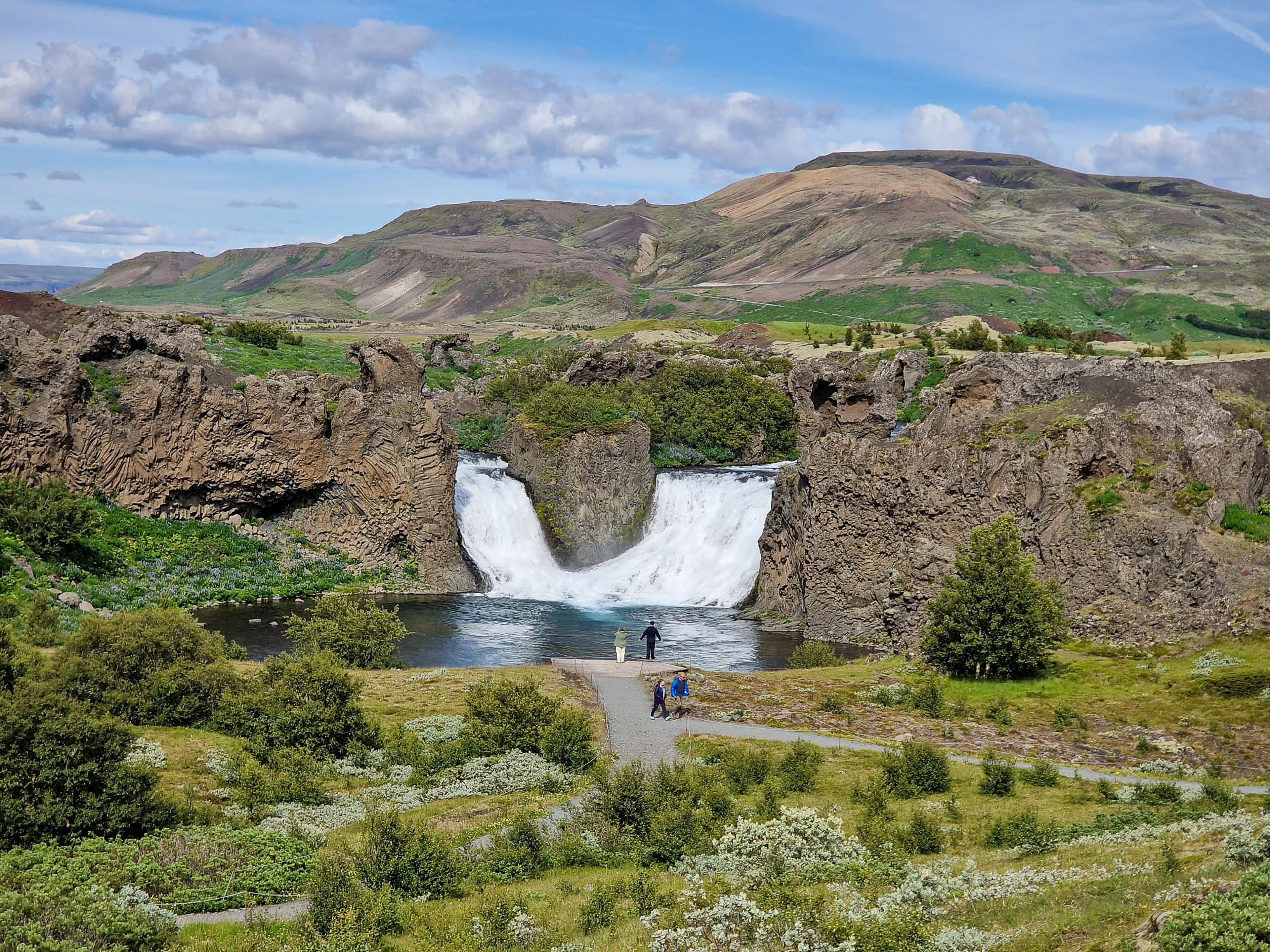 Two waterfalls plunge into a pool between rocky cliffs, set in a green, mountainous landscape with a few people.