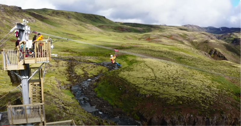 A person in a bright orange jacket and helmet is zip-lining across a lush, green valley with a small stream below. The launch platform on the left is occupied with more people in safety gear, waiting for their turn, set against a backdrop of rolling hills under a partly cloudy sky.
