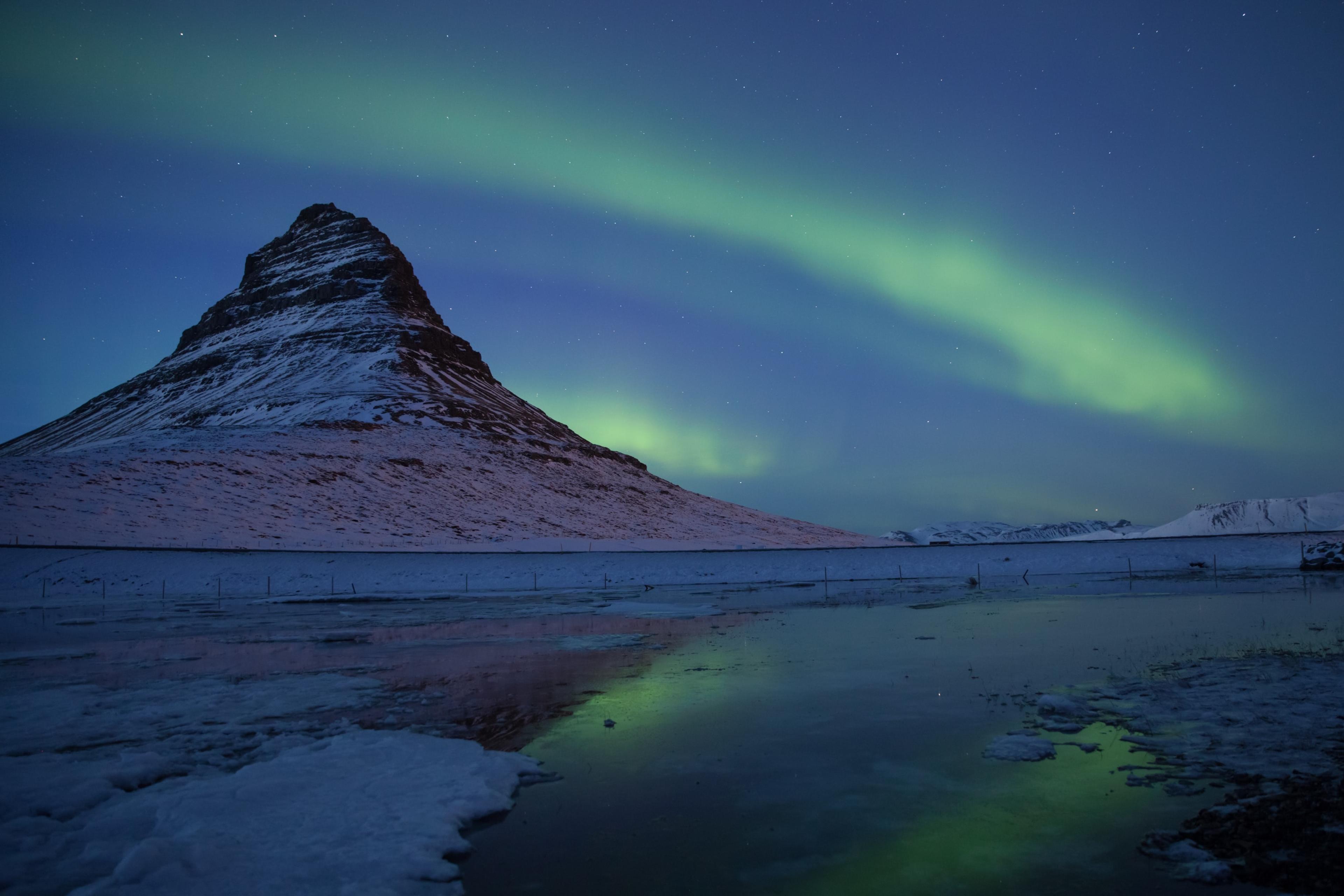 The Northern Lights arcing over Kirkjufell mountain and its reflection in a nearby water body during winter twilight.